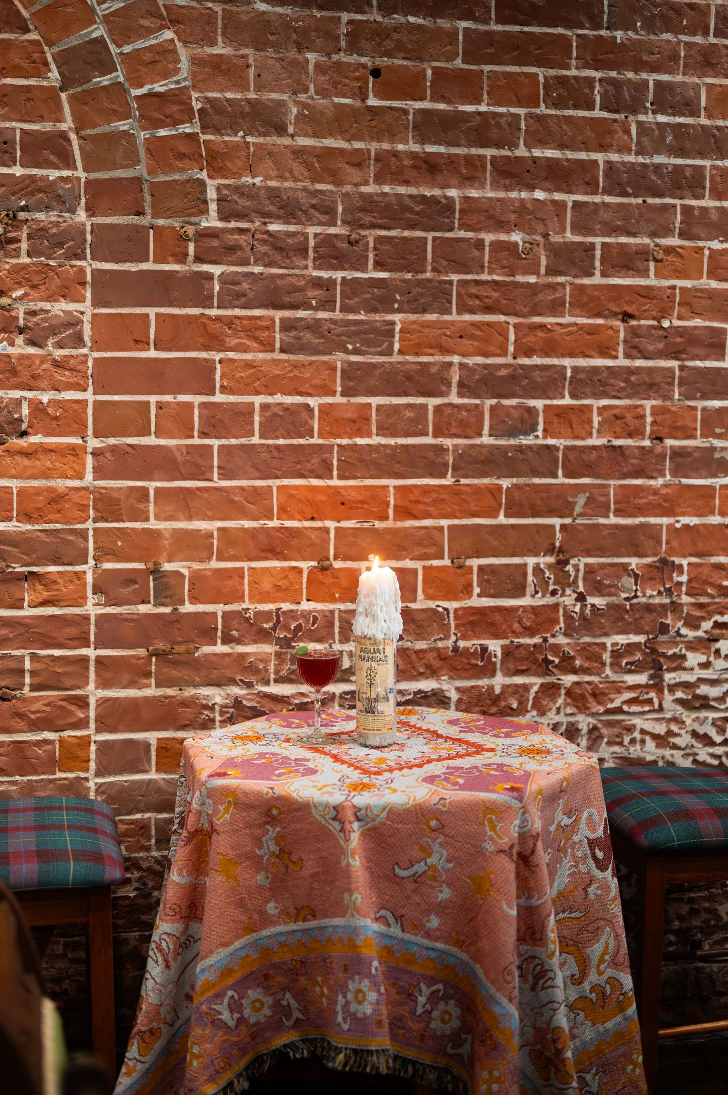 A lit candle and a glass of red wine sit on a round table with an ornate paisley cloth beside a rustic exposed brick wall.