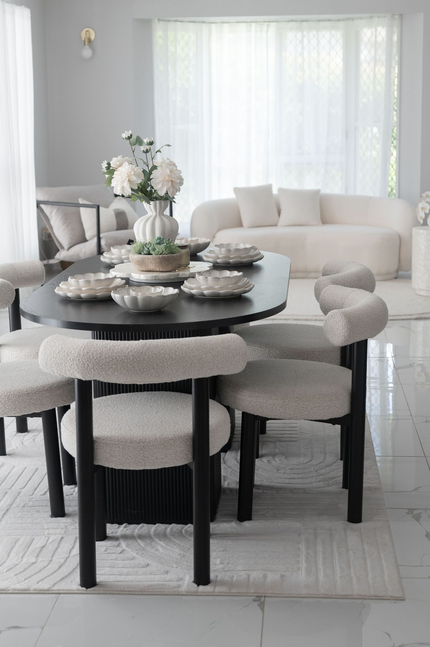 Modern dining room with a black oval table, boucle chairs, white floral centerpiece and a cream sofa near sheer curtains.