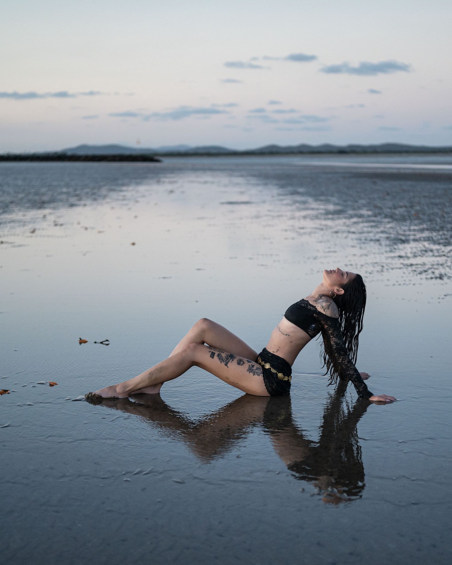 Woman in a black lace top and shorts reclines on a wet tidal flat, her reflection mirrored in the water at twilight.