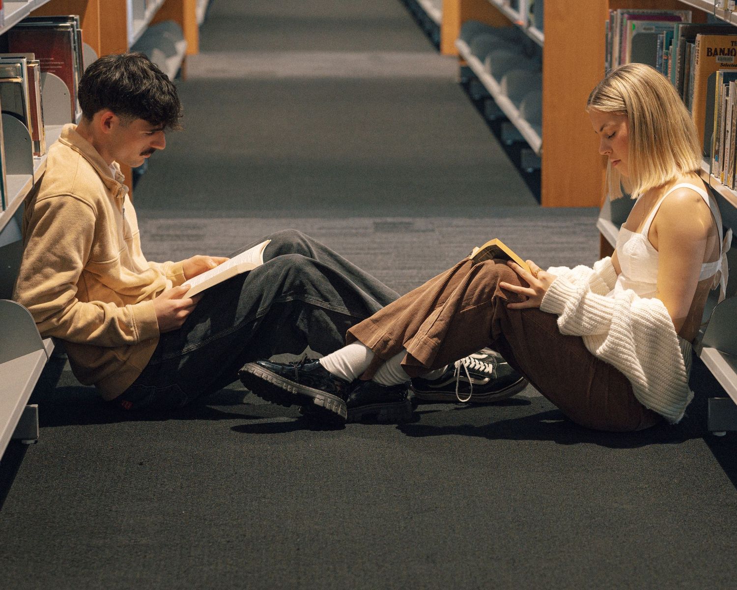 Man and woman sit on the library floor facing each other, each reading a book between rows of shelves.