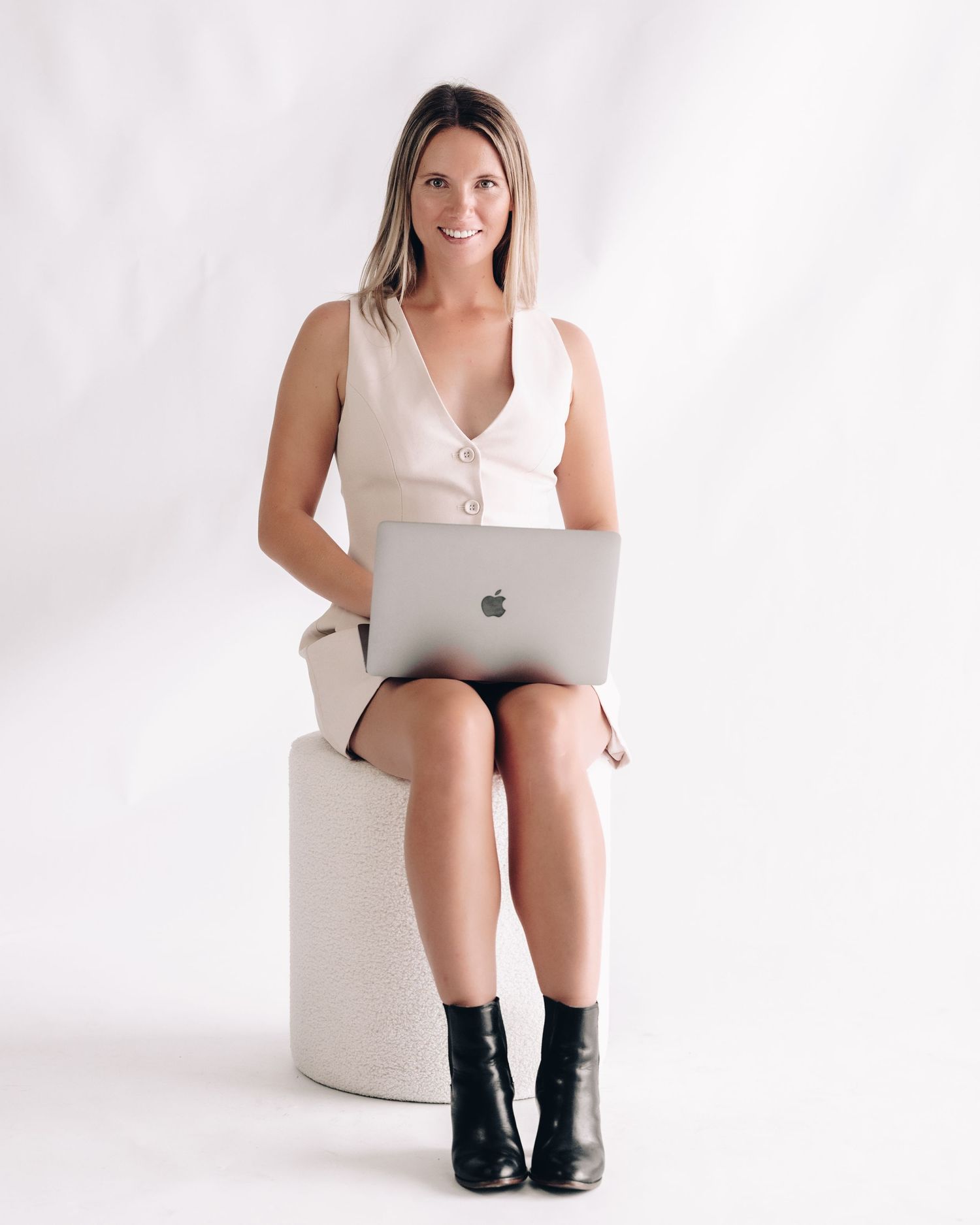 A smiling woman in a cream vest dress and black ankle boots sits on a white stool with a laptop on her knees.