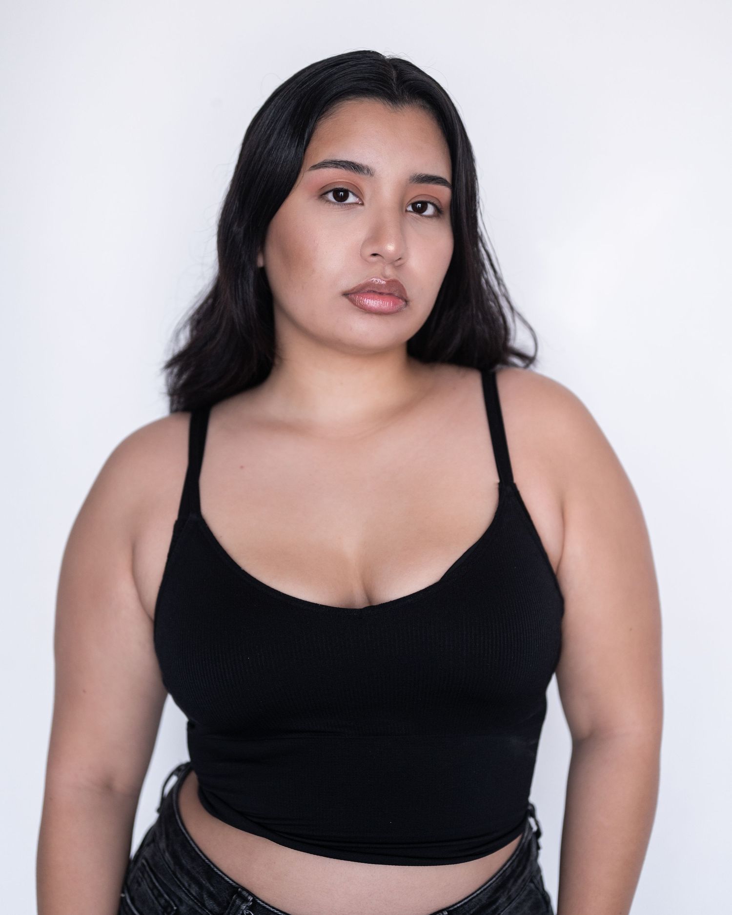 Portrait of a woman with long dark hair and natural makeup wearing a black cropped tank top against a plain white background.