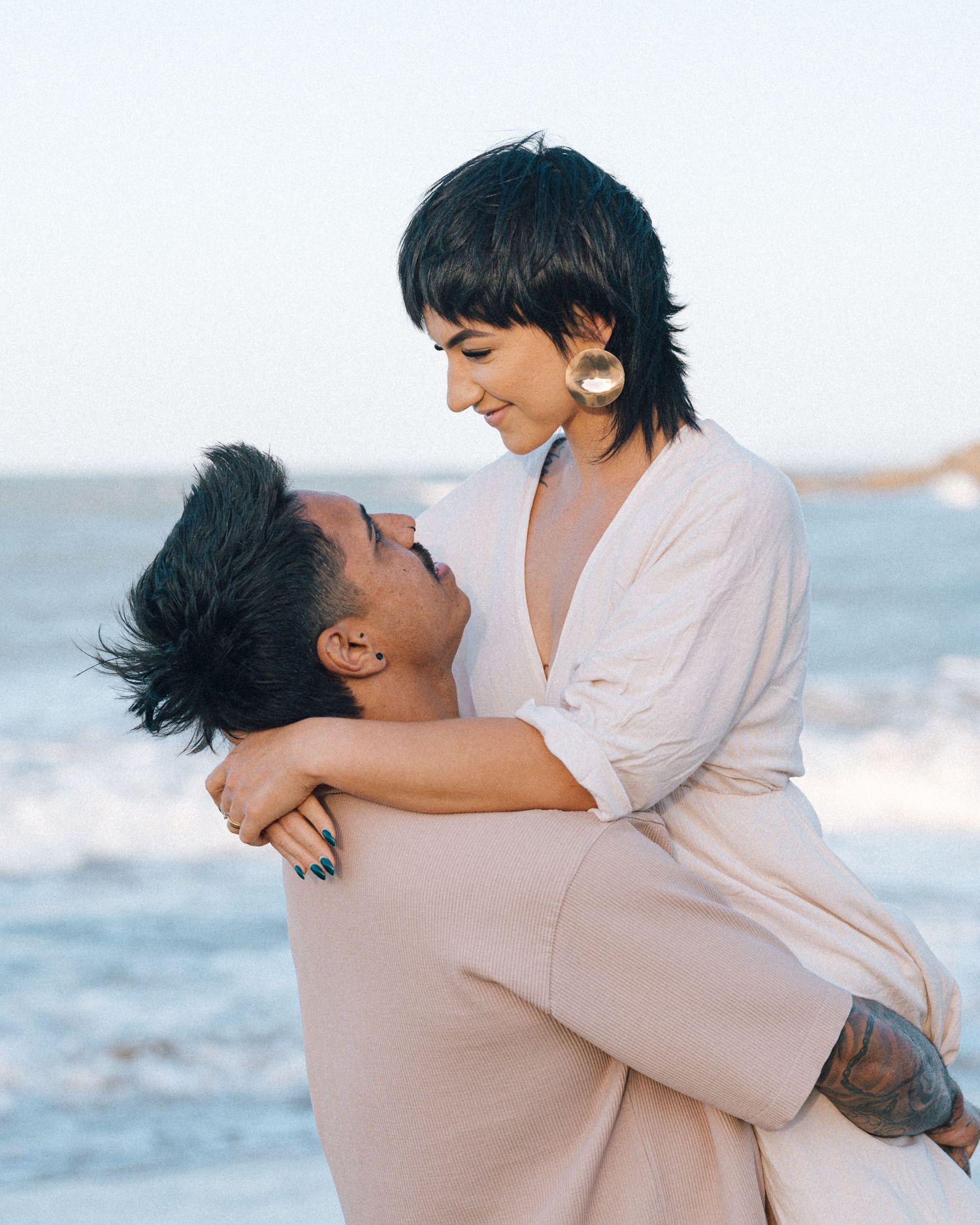 A smiling woman with a short dark hairstyle and gold earring is lifted in a joyful hug by a man on a sunlit beach.
