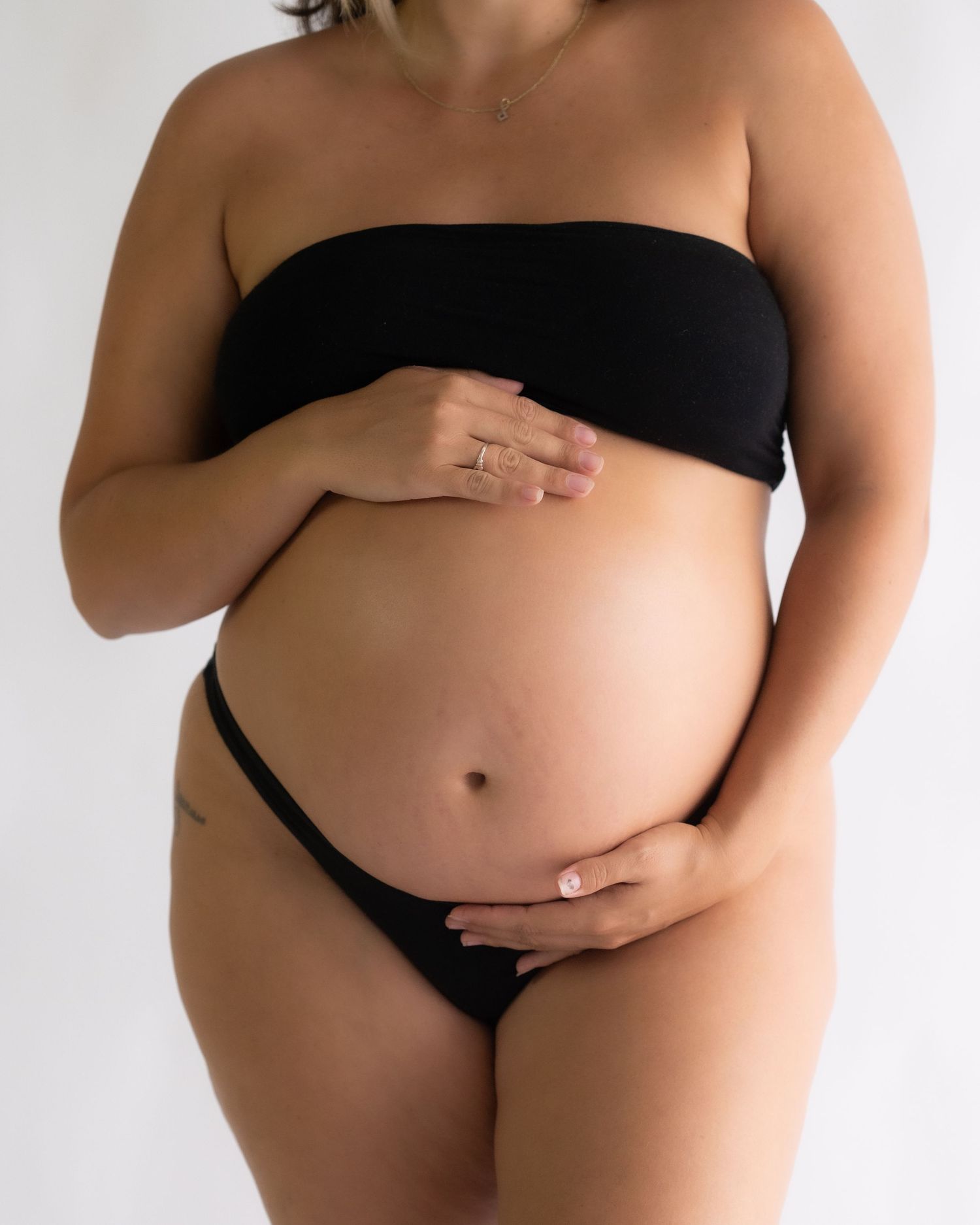Close-up maternity portrait of an expecting woman in a black bandeau top and briefs holding her belly with both hands.