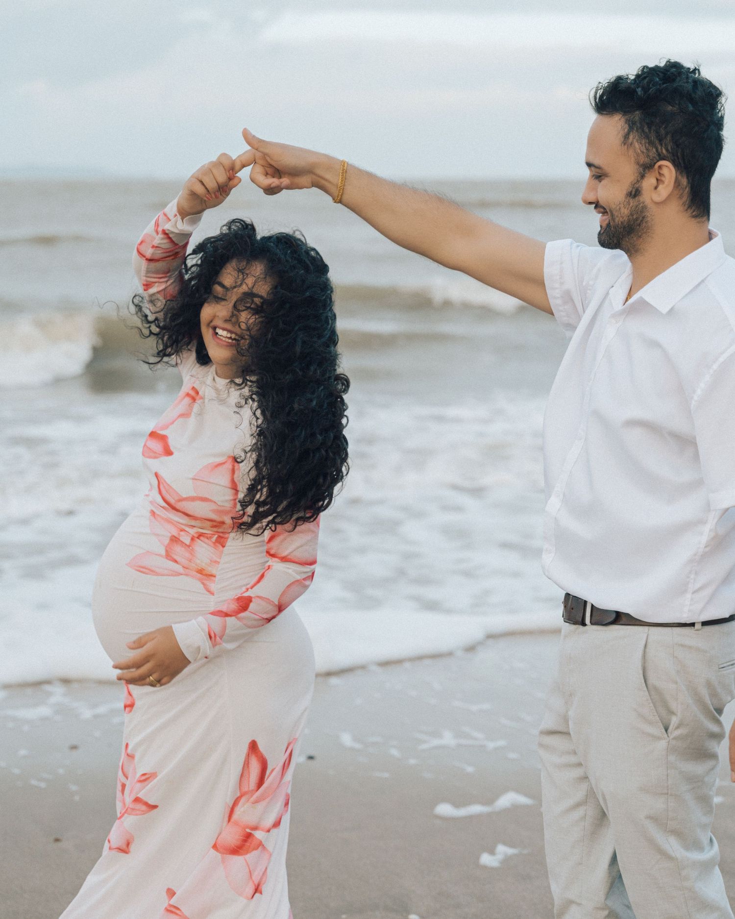Smiling pregnant woman twirls under her partner's raised arm as they dance together on the beach beside crashing waves.