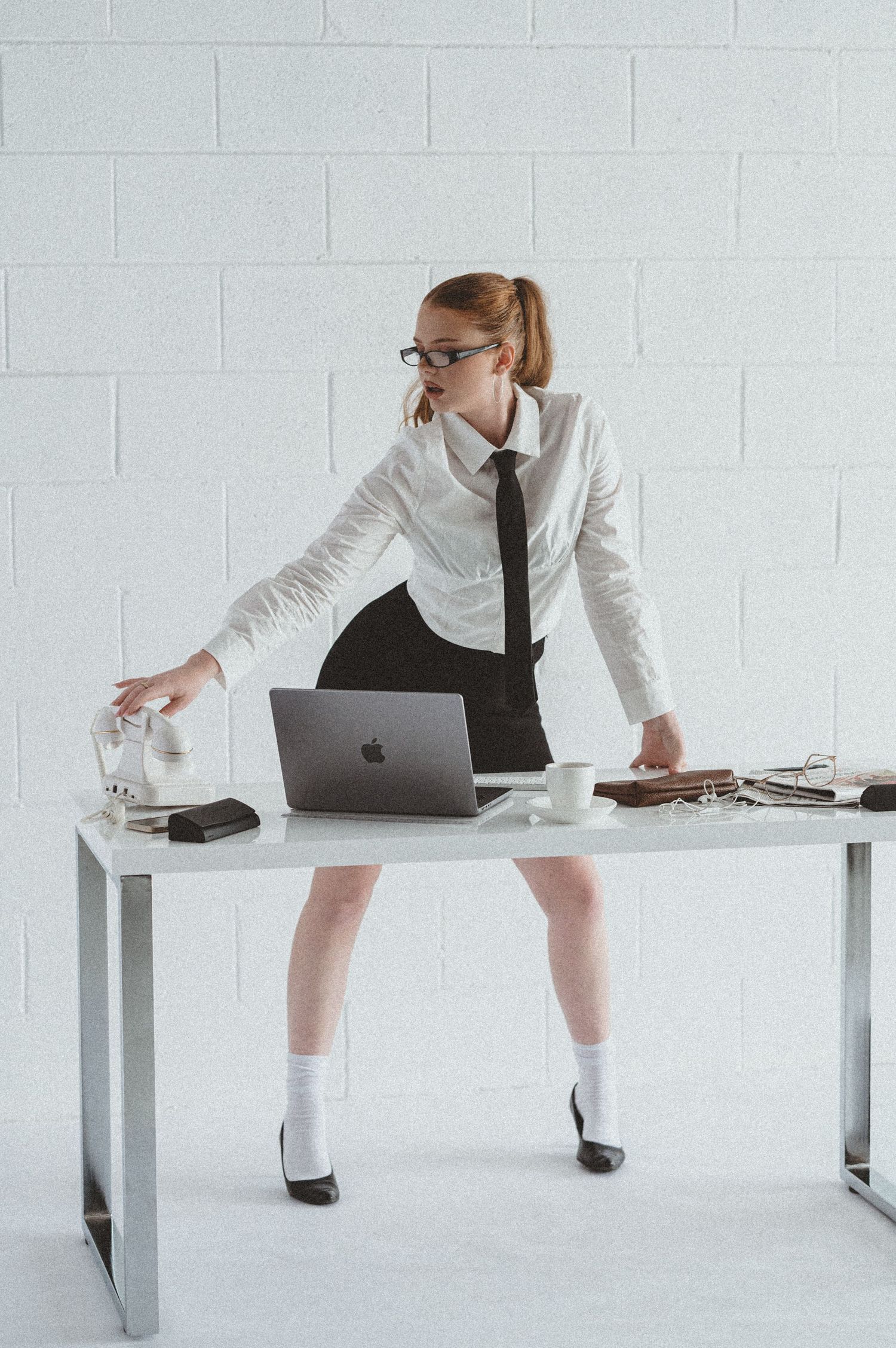 Woman in white shirt, black tie and skirt with glasses leaning over a modern desk with a laptop in a bright office.
