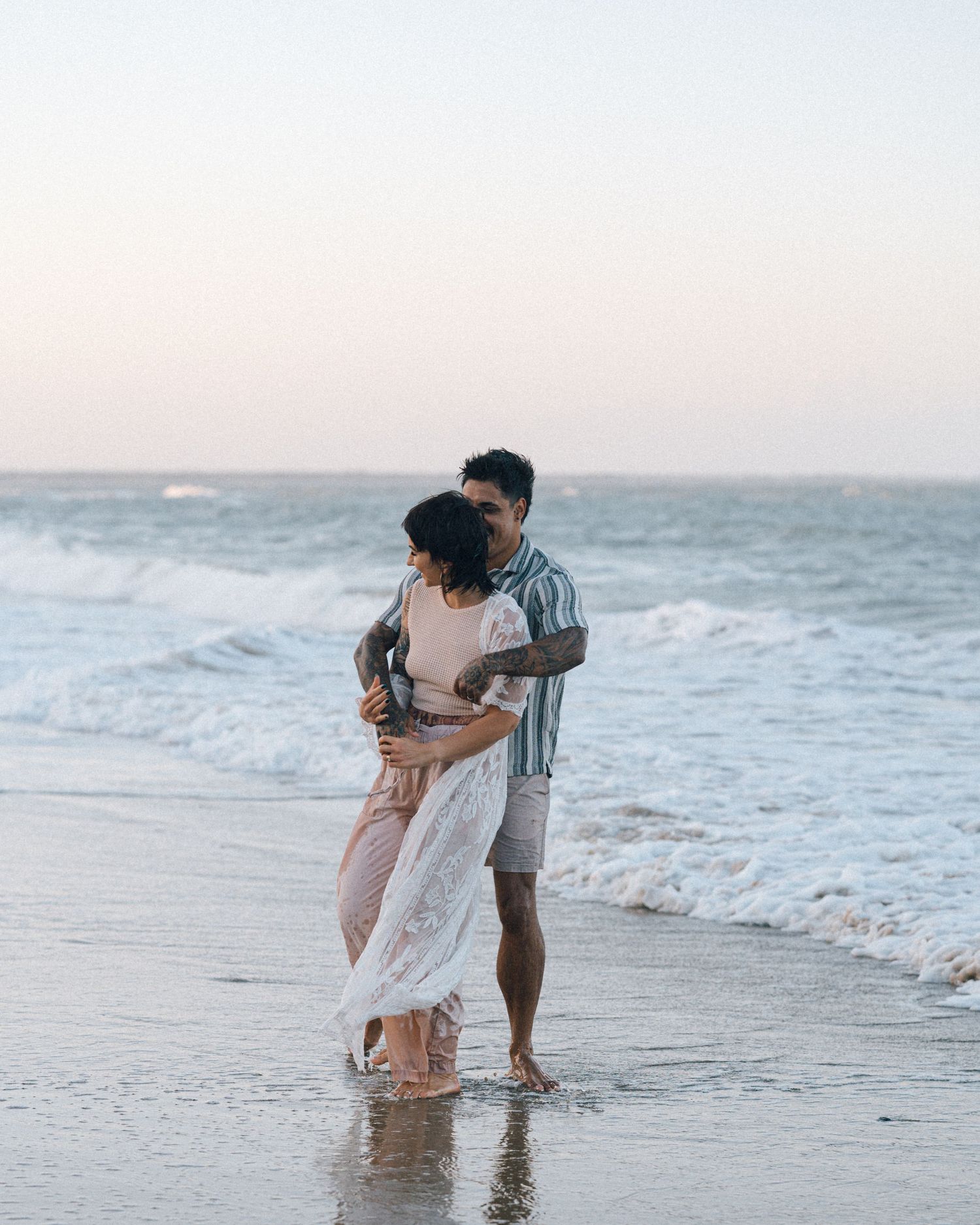 Couple embracing in the shallow ocean waves at sunset, the woman in a white lace dress and the man in a striped shirt.