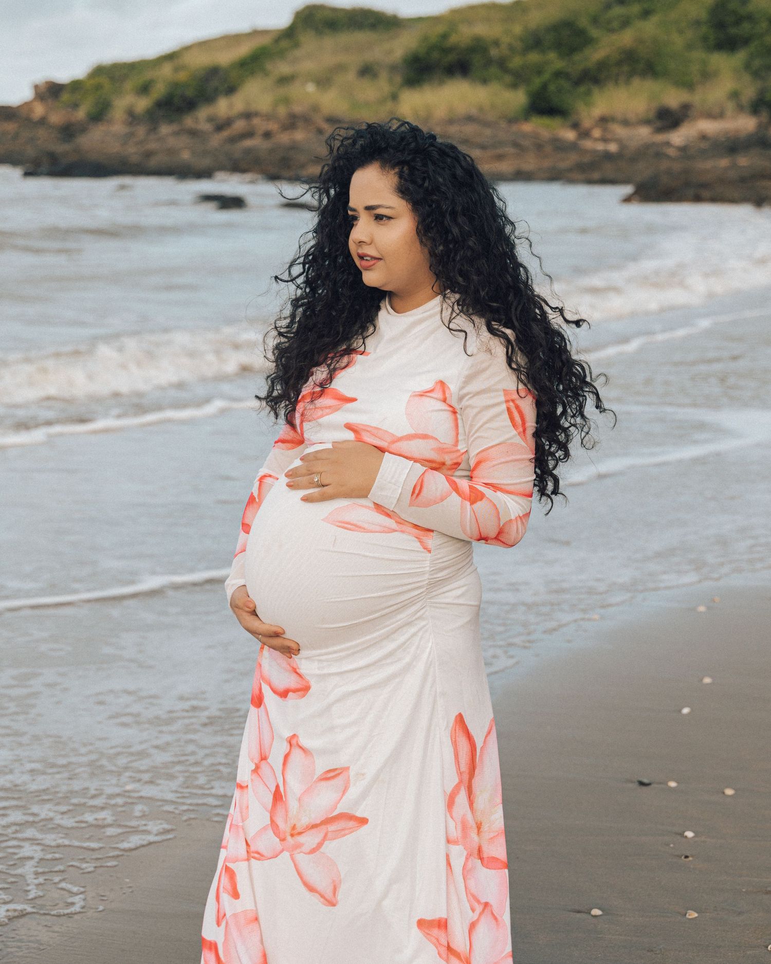 Expecting mother in a floral maternity dress cradles her baby bump while gazing out at the waves on a rocky shoreline.