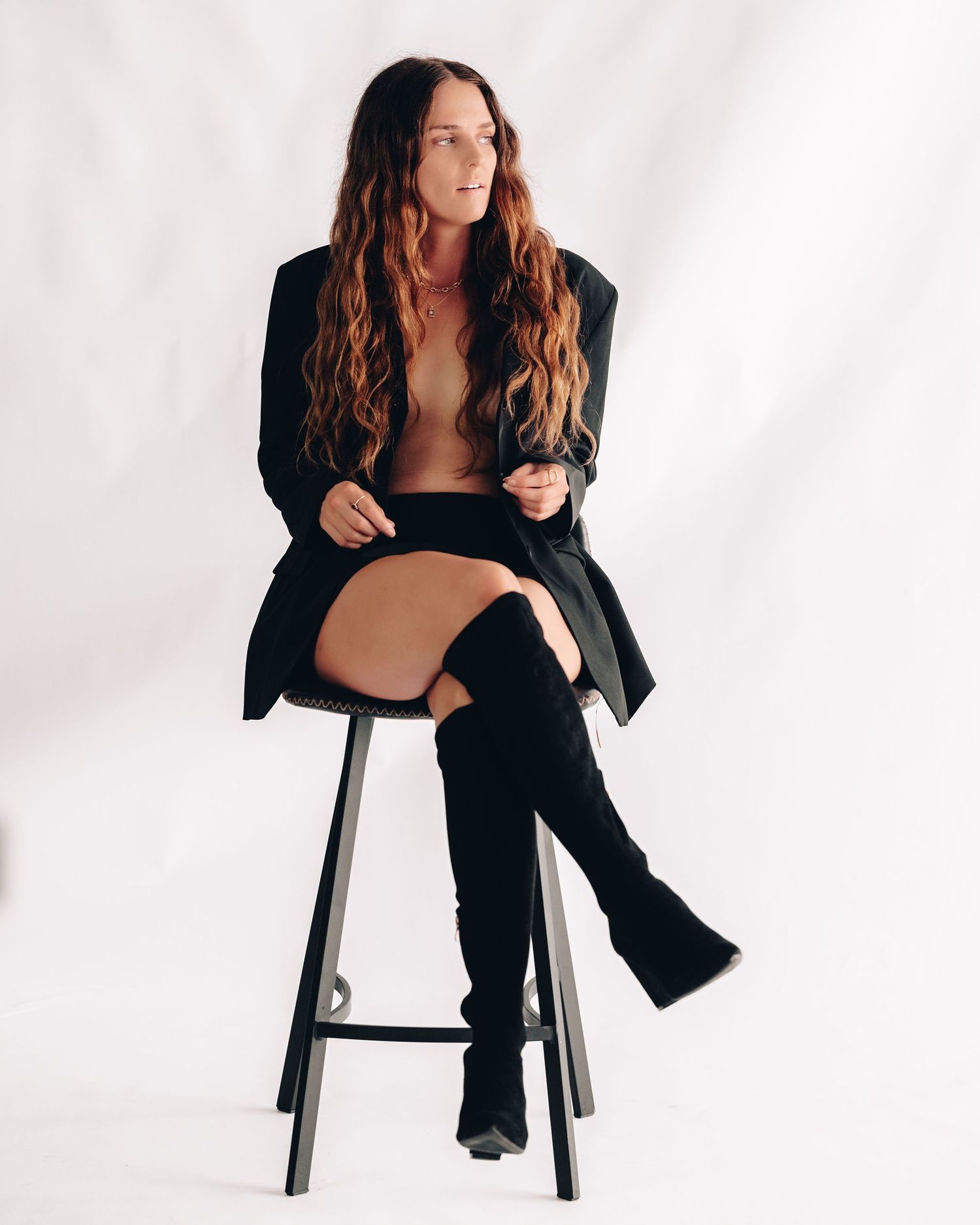 Woman with long wavy hair in a black blazer and thigh-high boots sits on a black stool, looking away from the camera.