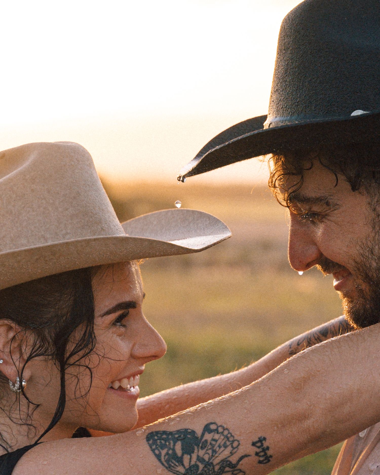 A smiling couple in cowboy hats gaze at each other at sunset, the woman's tattooed arm wrapped around the man.