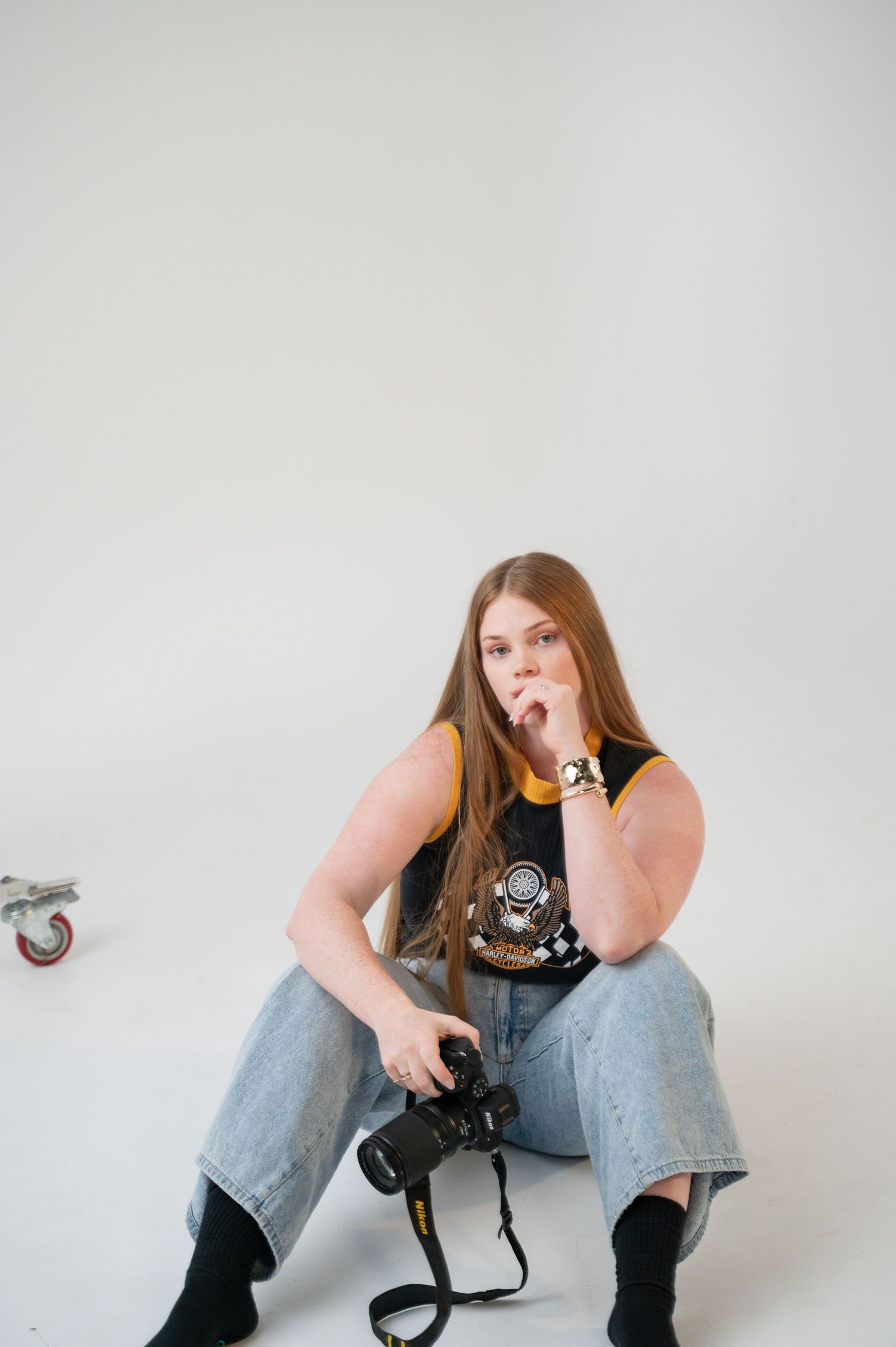 Woman with long auburn hair sits on a white studio floor in light jeans and a graphic tank top, holding a camera and gazing at the viewer.