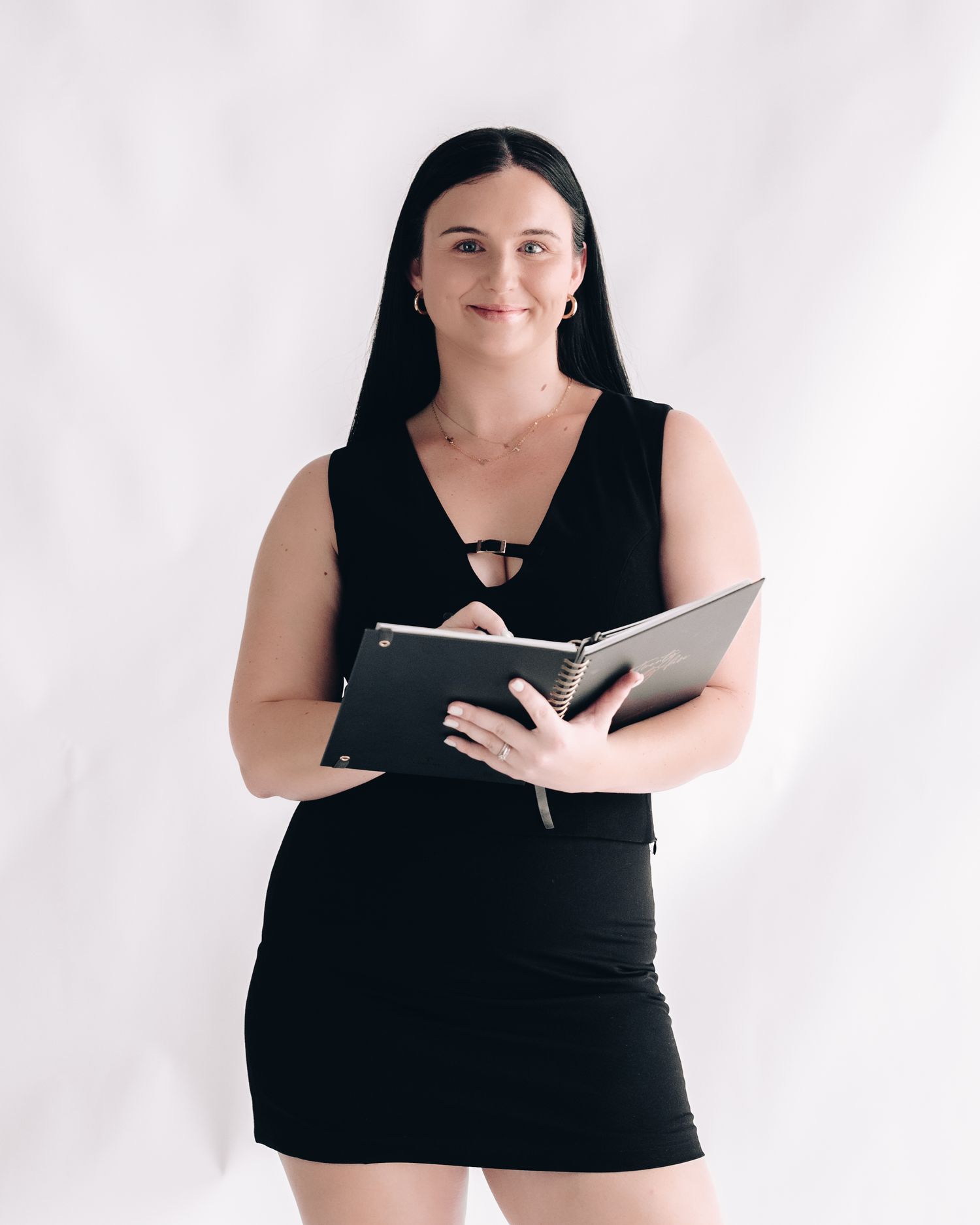 Smiling woman with long dark hair in a black dress holds an open notebook while standing against a plain white backdrop.