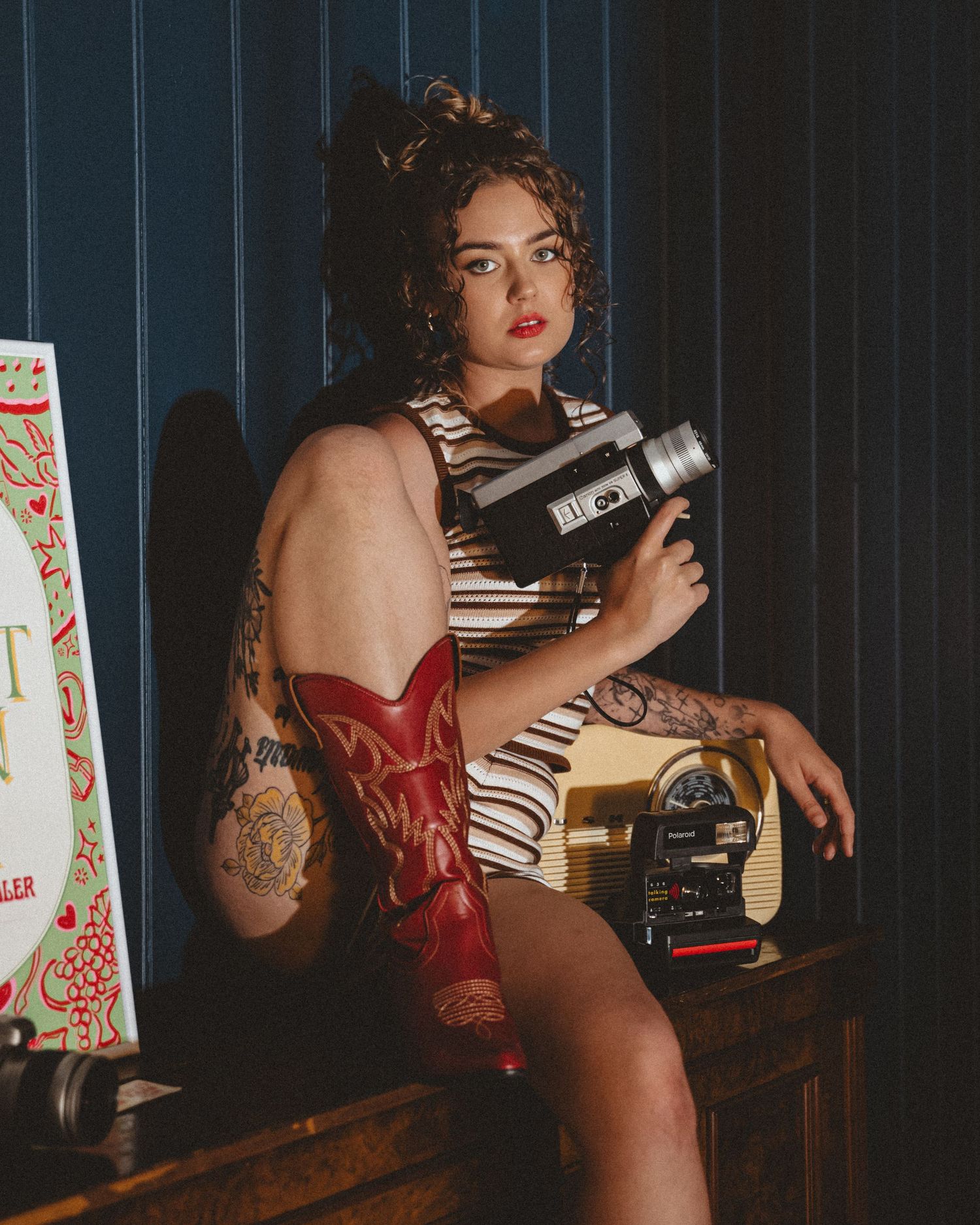 Tattooed woman with curly hair and red lipstick wears a red cowboy boot and holds a vintage film camera while seated on a wooden cabinet.
