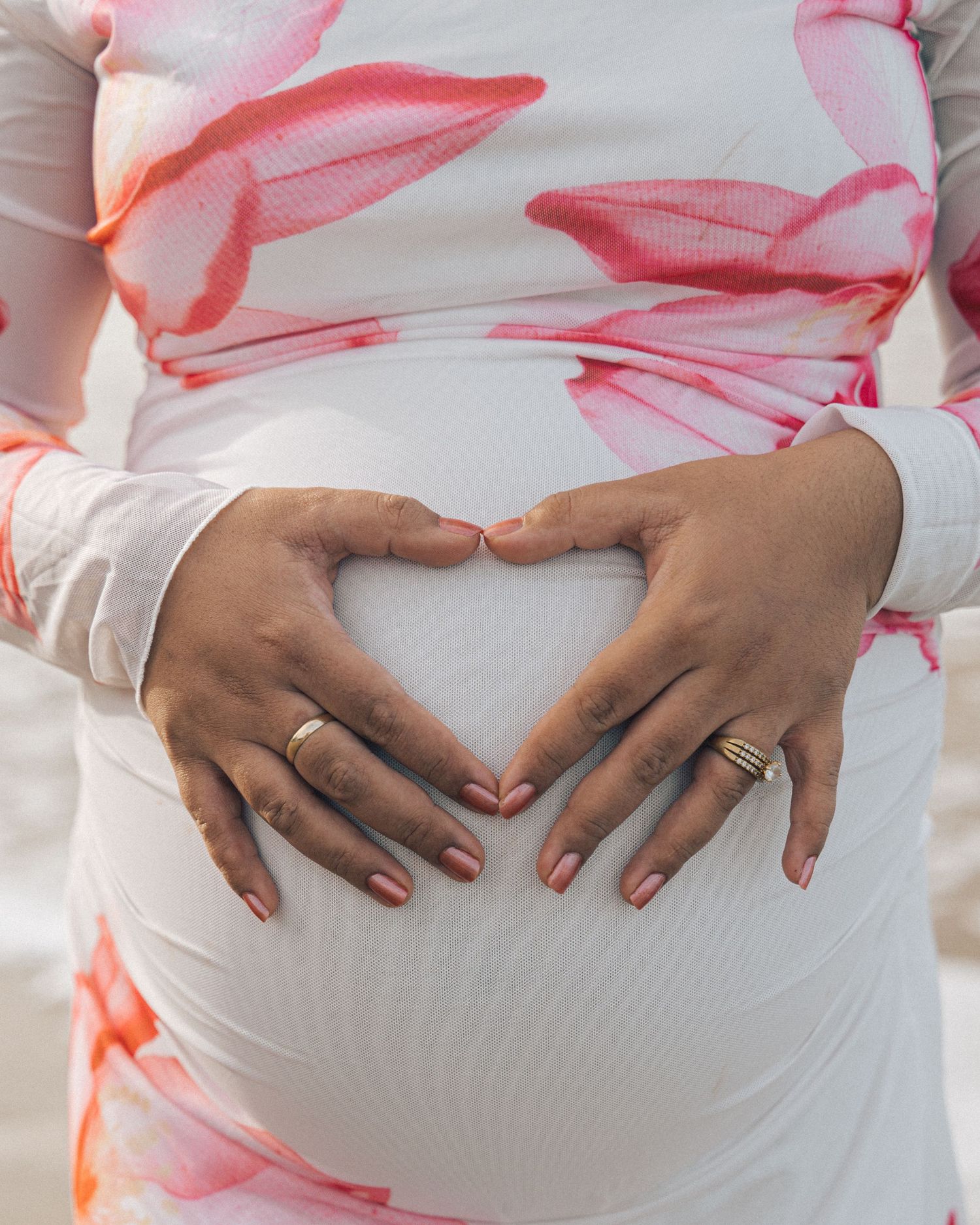 Close-up of a pregnant woman's hands forming a heart shape on her belly, wearing rings and a pink floral maternity dress.