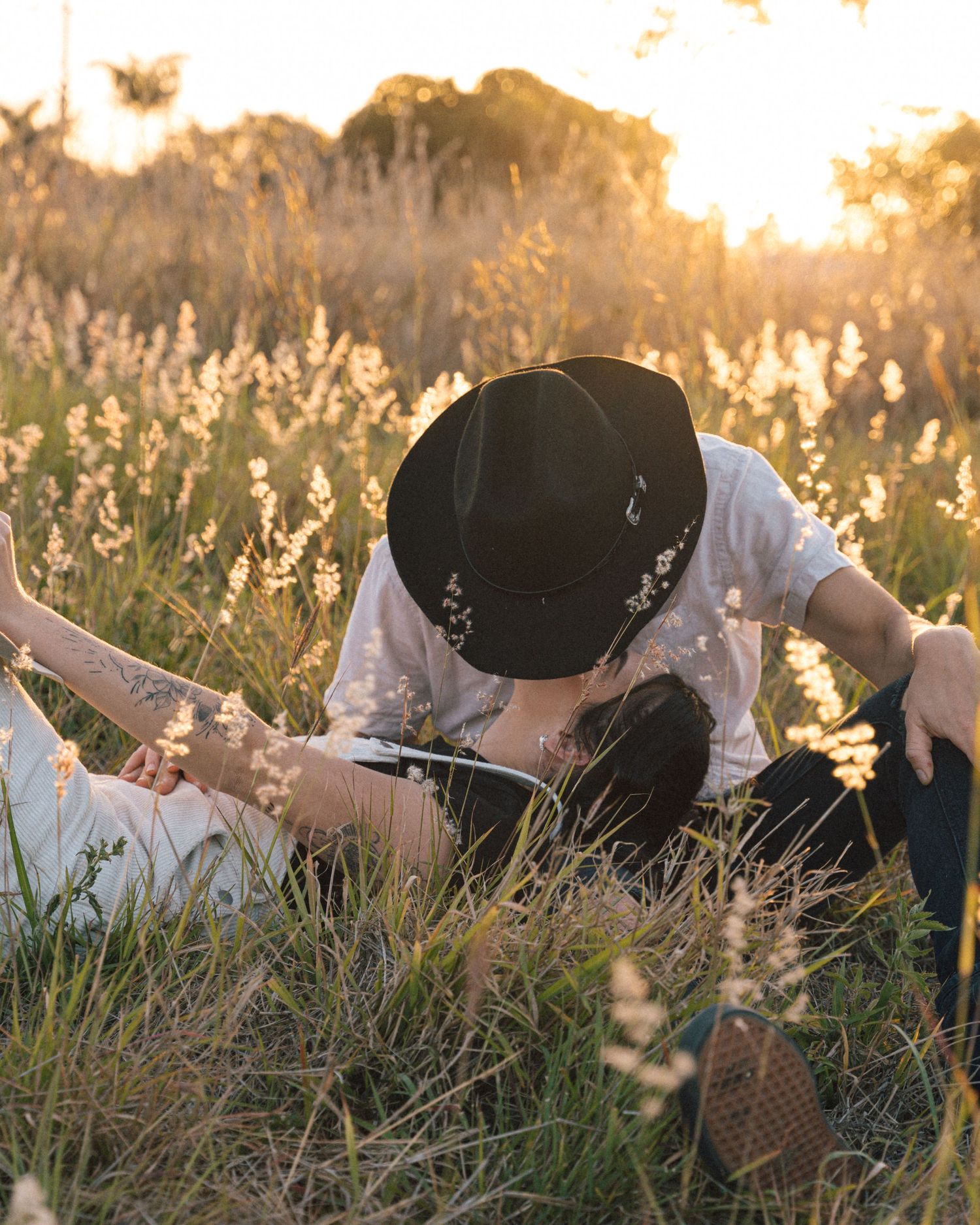 Couple in a black hat and white shirt lie in tall wildflower grass at golden hour, sharing an intimate moment.