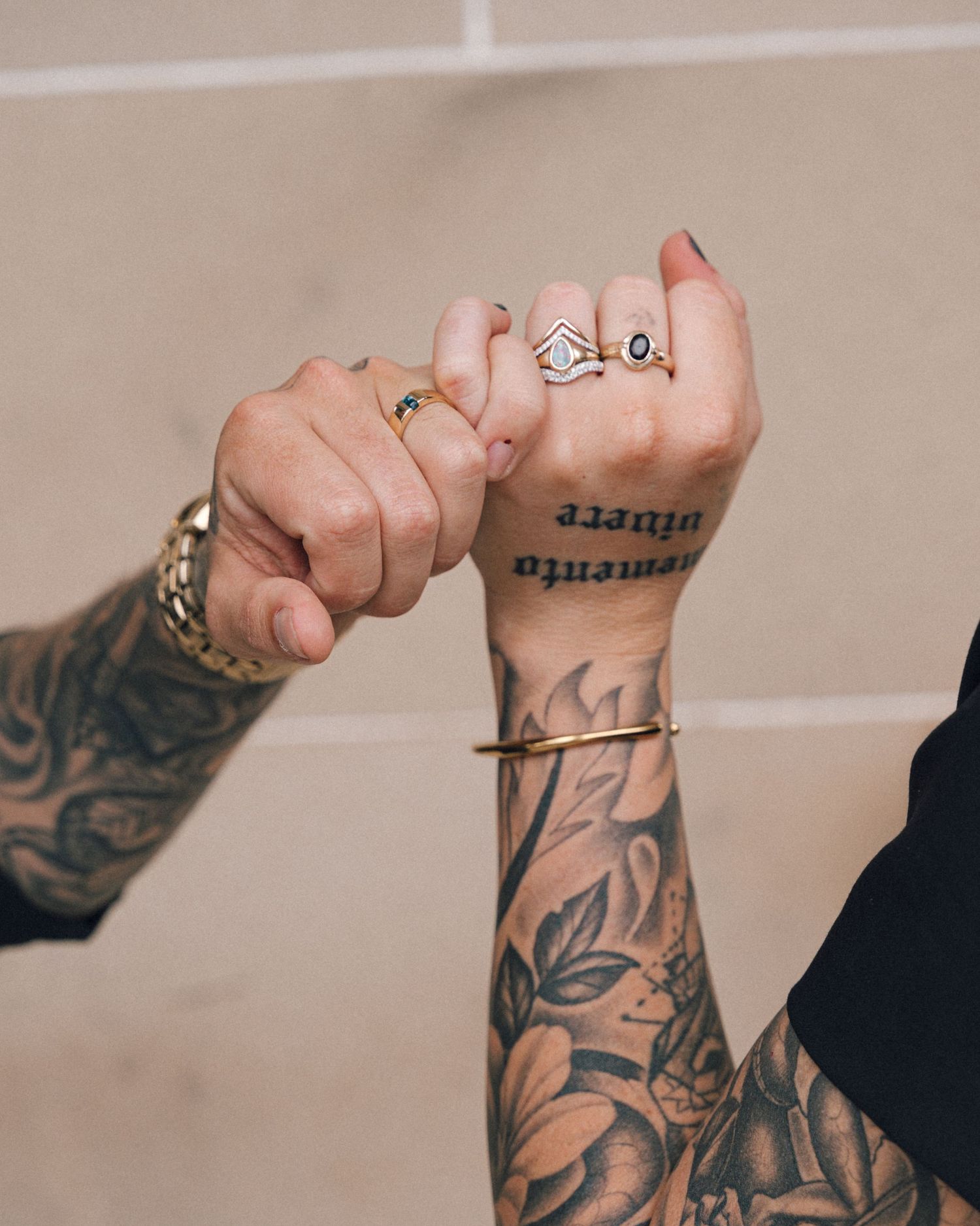 Two tattooed hands with pinky fingers linked, showing stacked gold rings and a floral forearm tattoo with the words memento vivere.