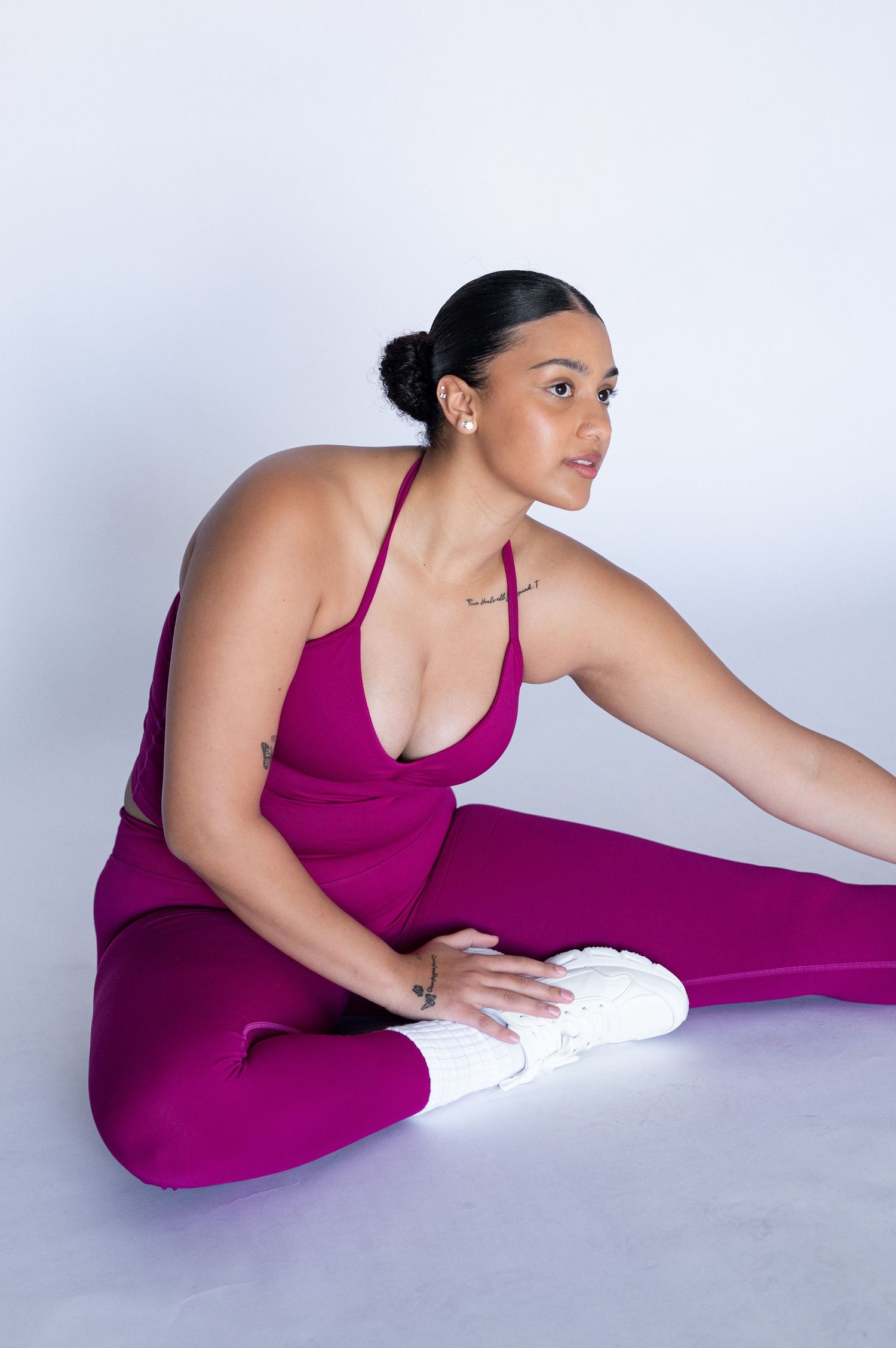 Woman in a magenta athletic set and white sneakers sits on the floor stretching her leg against a plain light background.