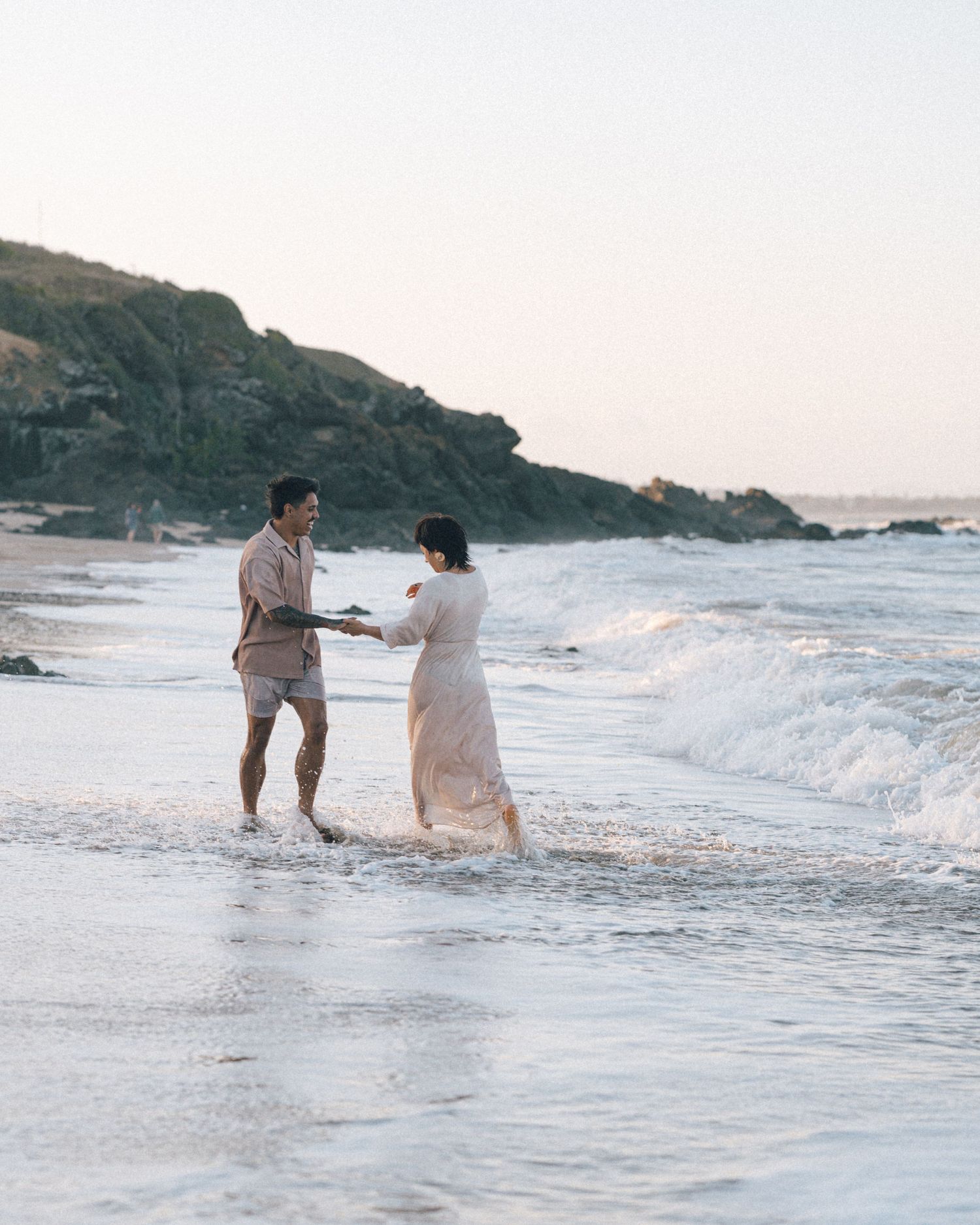 A couple in light clothing holds hands and plays in shallow ocean waves near a rocky coastal headland at sunset.