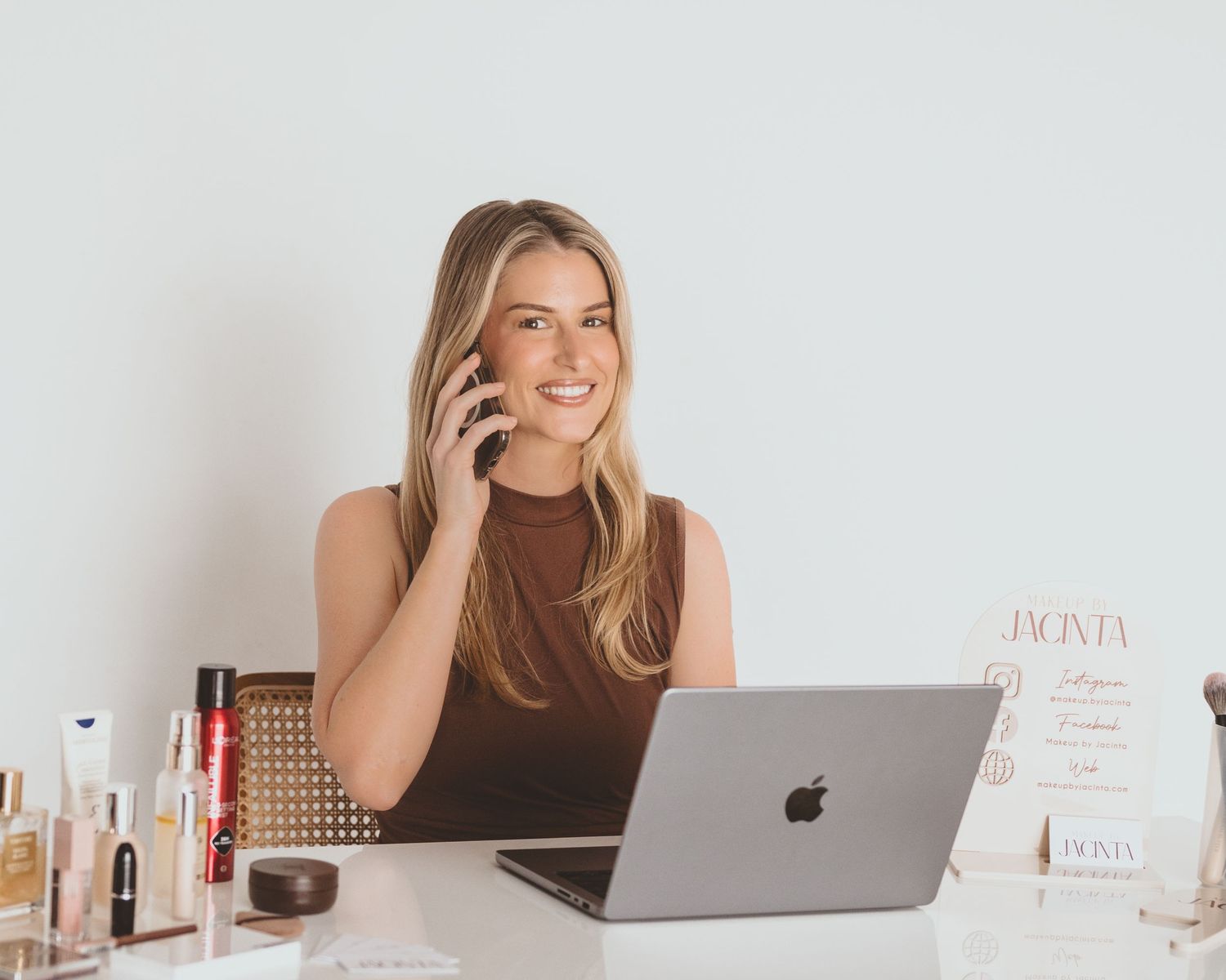 Smiling woman in a brown top talks on the phone at a white desk with a laptop and skincare and makeup products beside her.