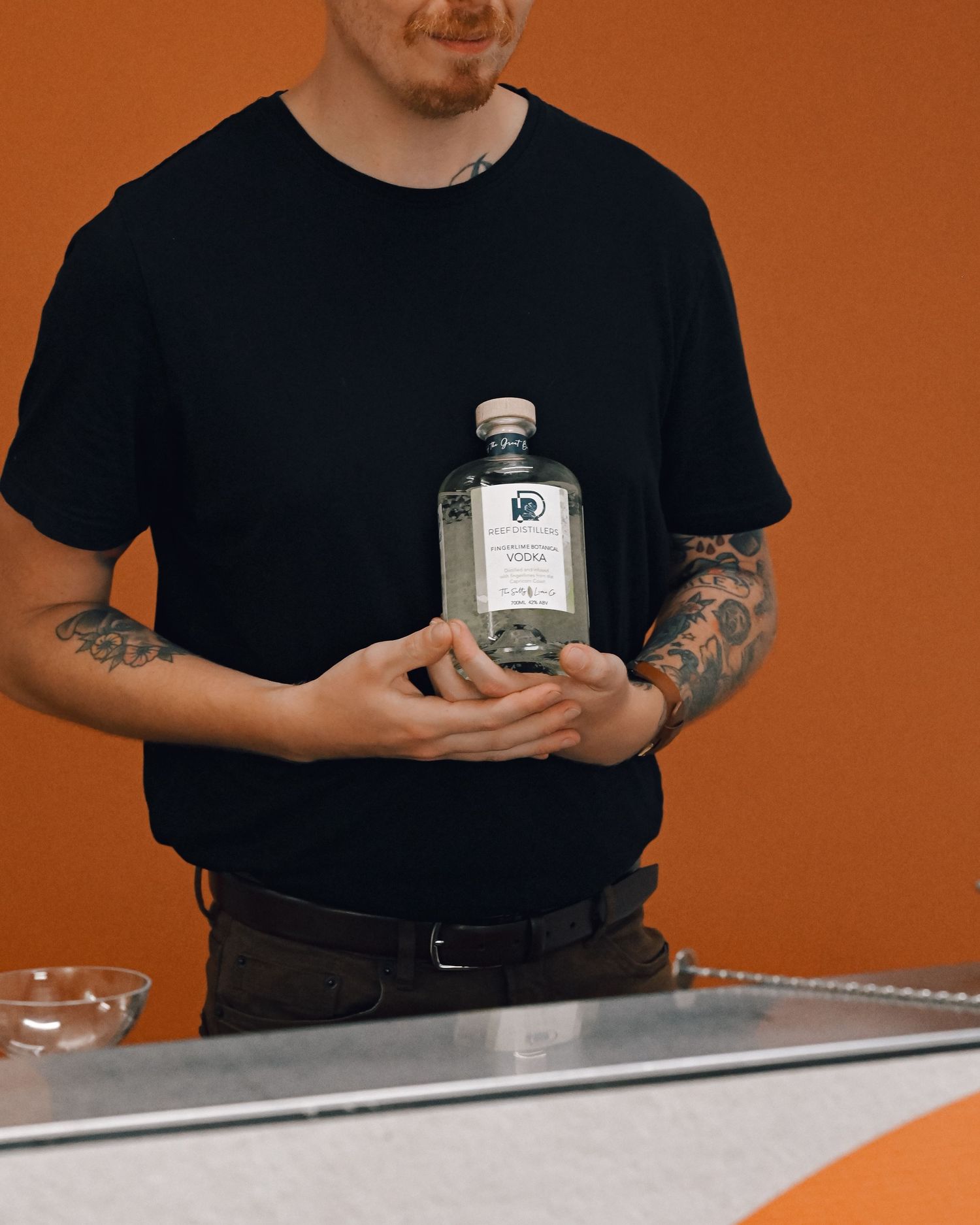 Smiling tattooed bartender in a black tee holds a clear bottle of vodka in front of a bold orange wall.