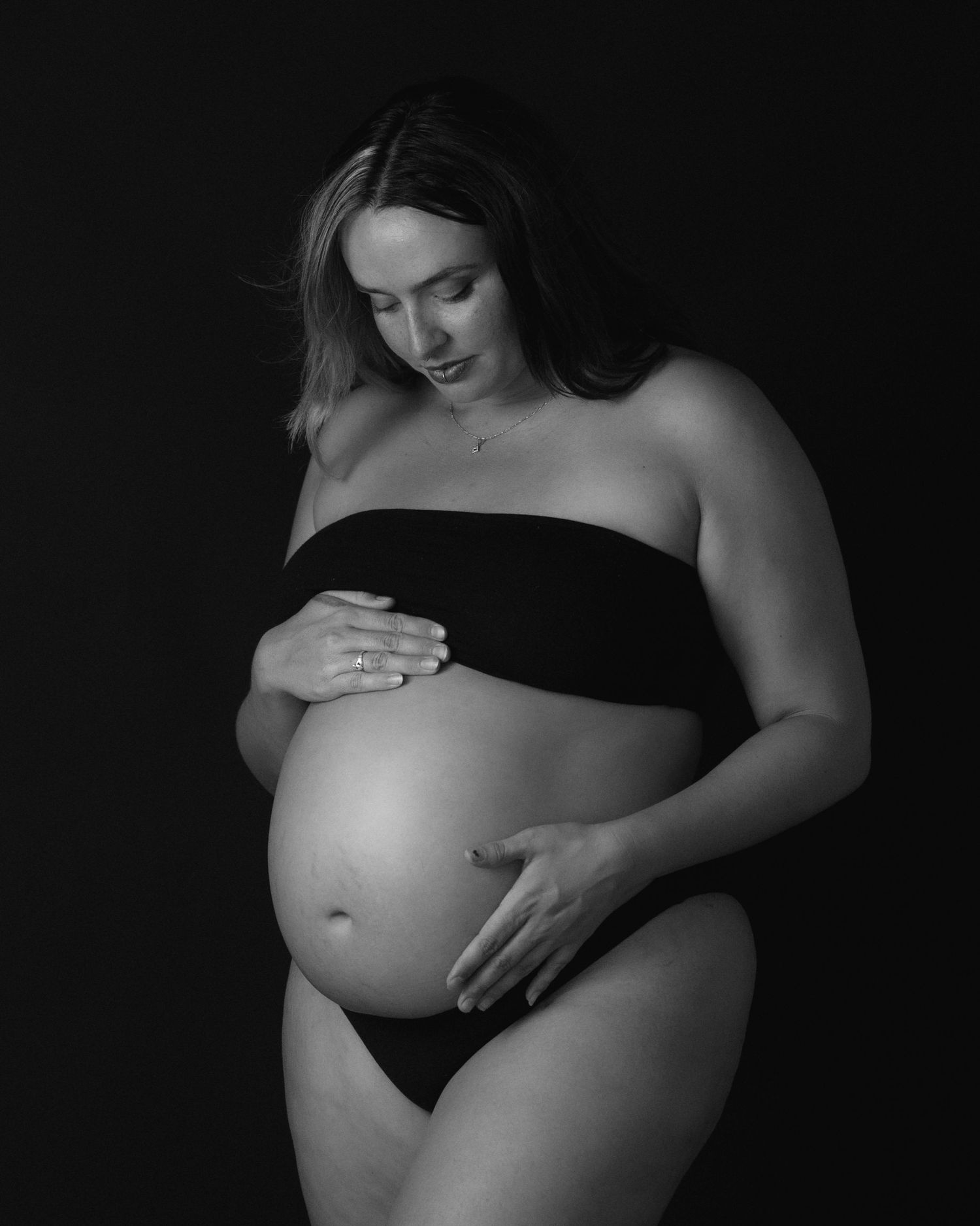Black and white studio maternity portrait of a pregnant woman looking down and cradling her belly in a strapless top.