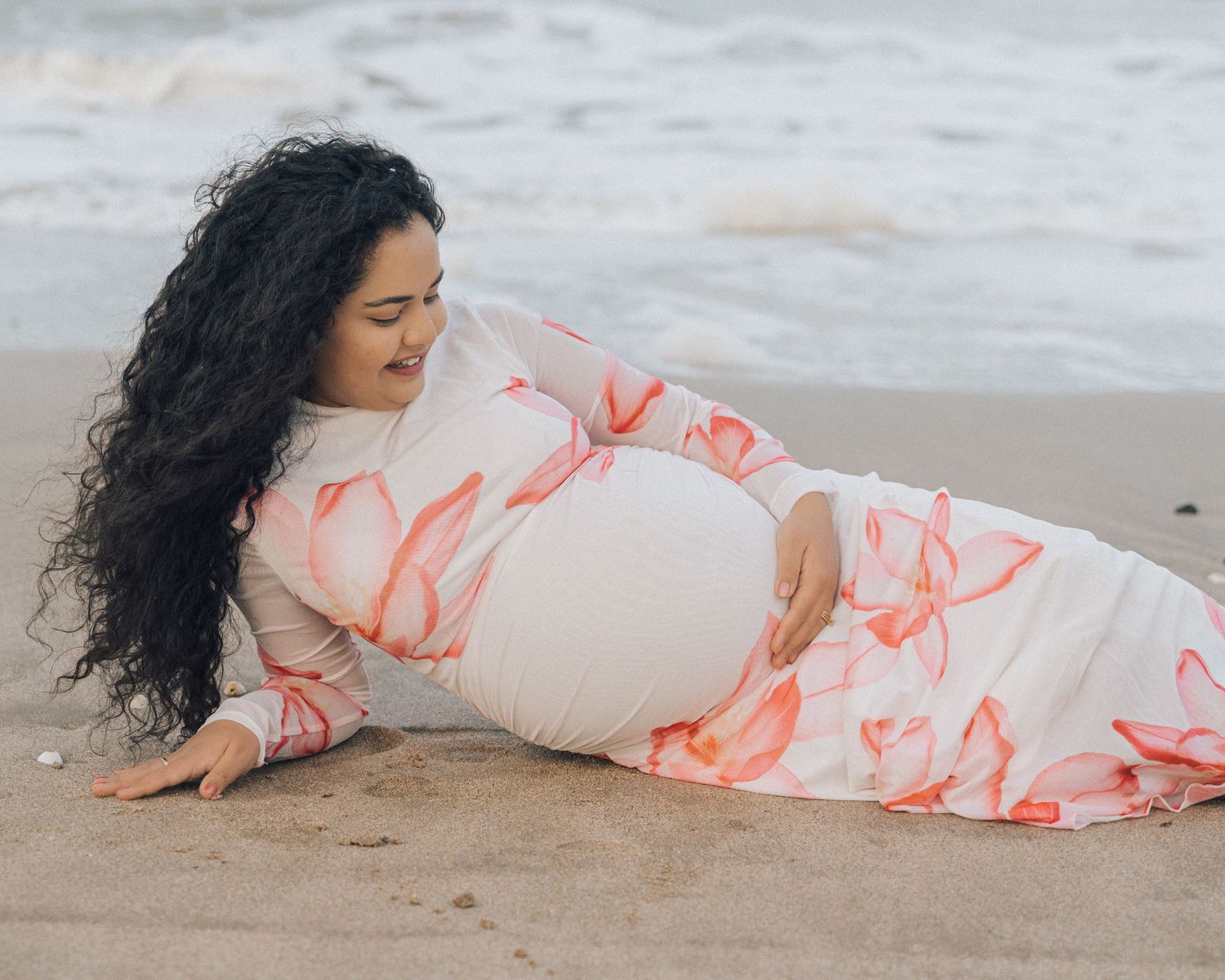 Pregnant woman in a pink floral dress lies on her side on the sand, smiling and holding her belly near the ocean.