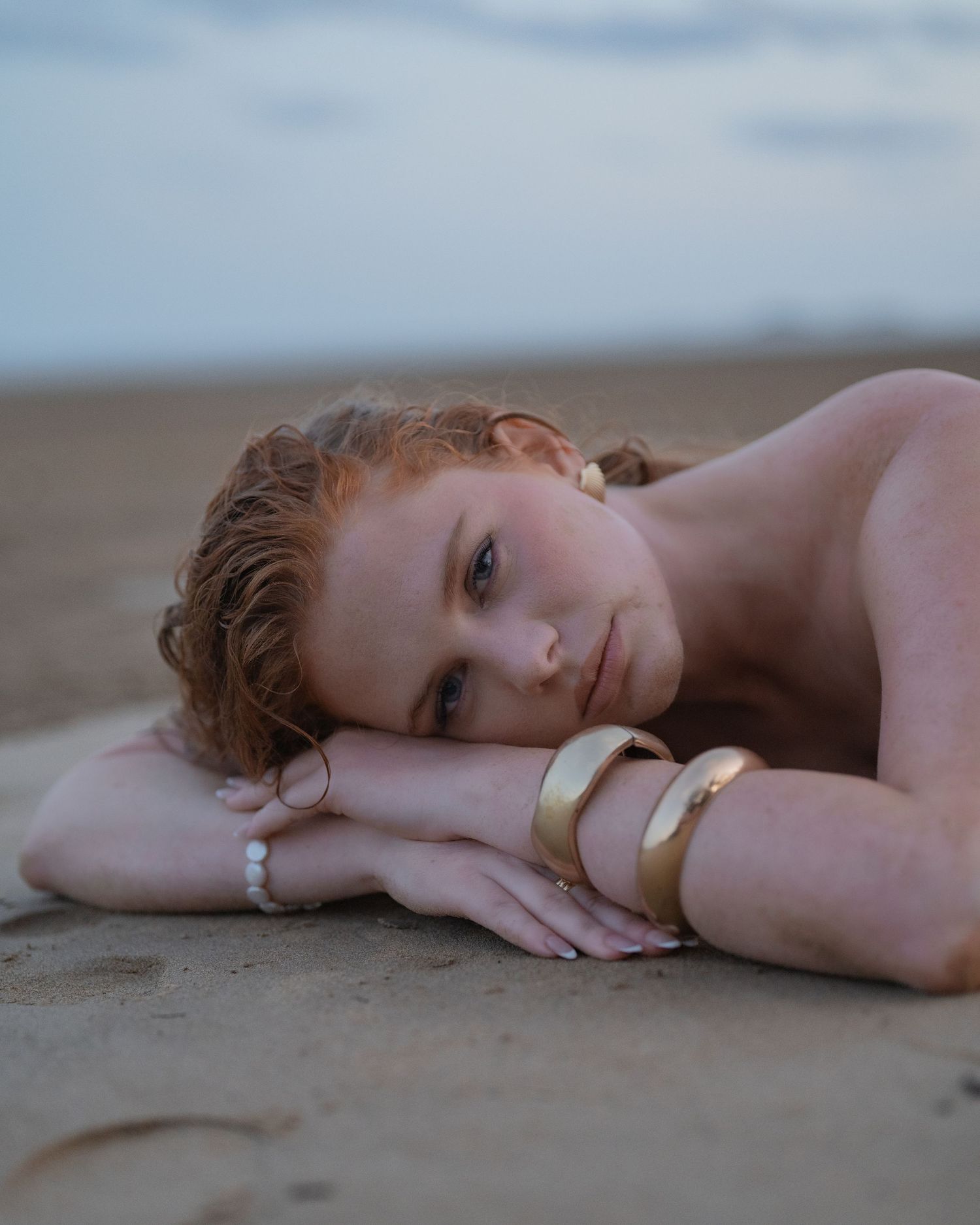 Red-haired woman with wet hair rests her head on her arms on a sandy beach, wearing chunky gold bangles at dusk.