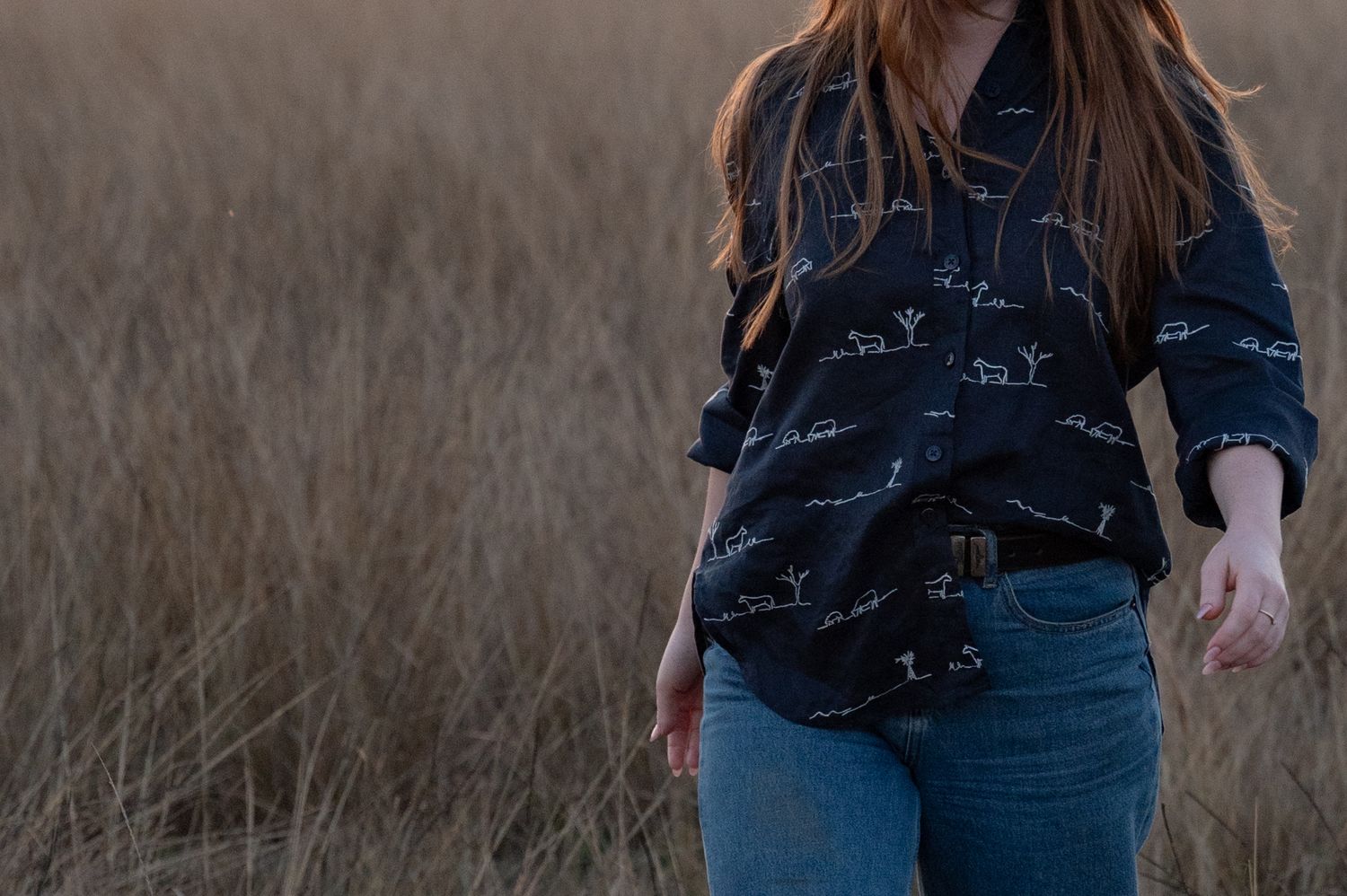 Woman with long red hair walking through tall dry grass in a navy button-up shirt with a printed pattern and blue jeans.