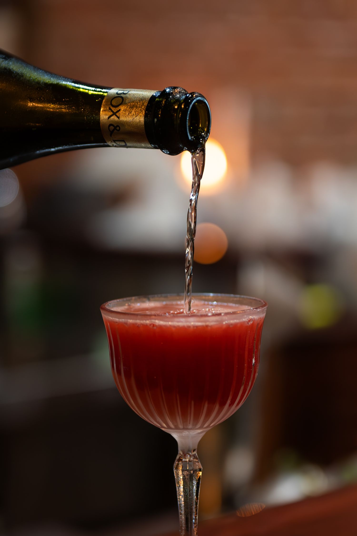 Bartender pours sparkling wine from a bottle into a coupe glass filled with a vibrant red cocktail at a bar.