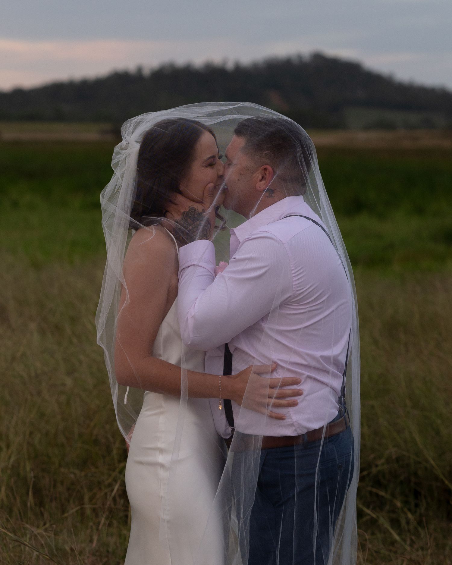 Bride and groom share a kiss beneath a sheer wedding veil in an open field with hills behind.