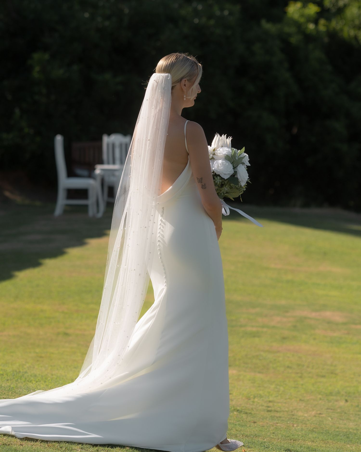 Bride in a satin wedding gown with a long veil and white bouquet stands on a sunny garden lawn near white chairs.