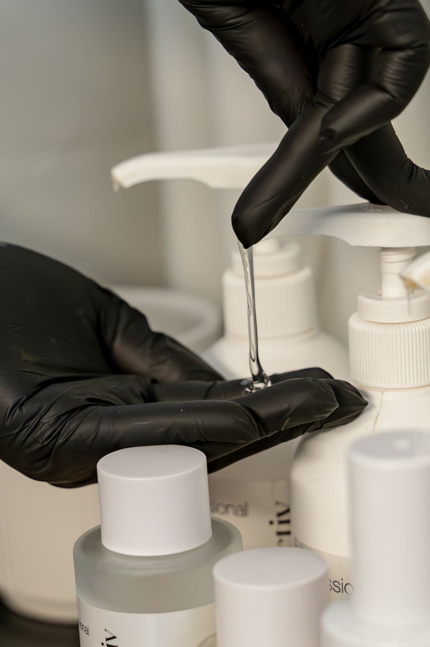 Black-gloved hands dispense clear liquid from a pump bottle onto a gloved palm, surrounded by white skincare product bottles.