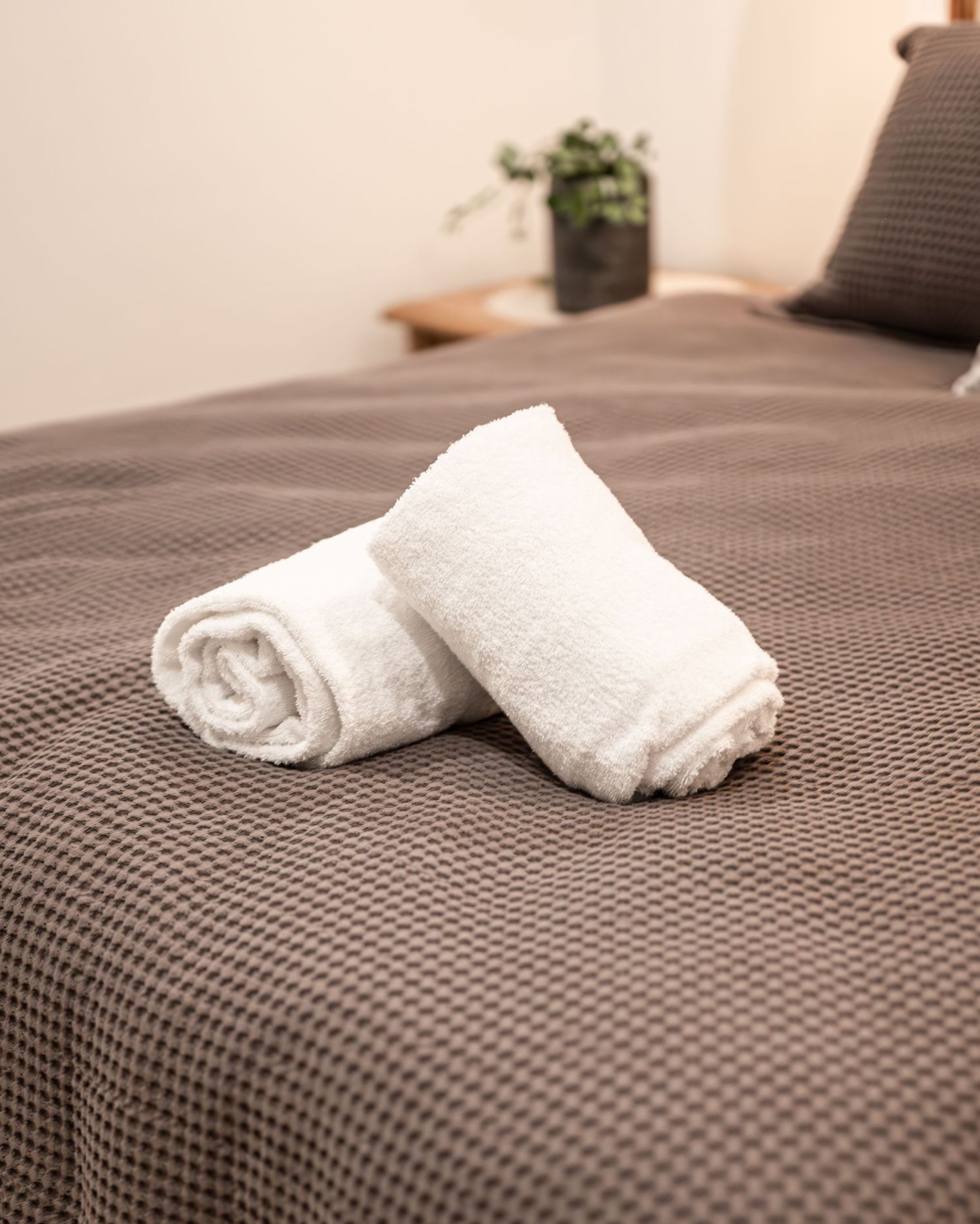 Two neatly rolled white towels resting on a taupe waffle-weave bedspread in a tidy, minimalist bedroom.
