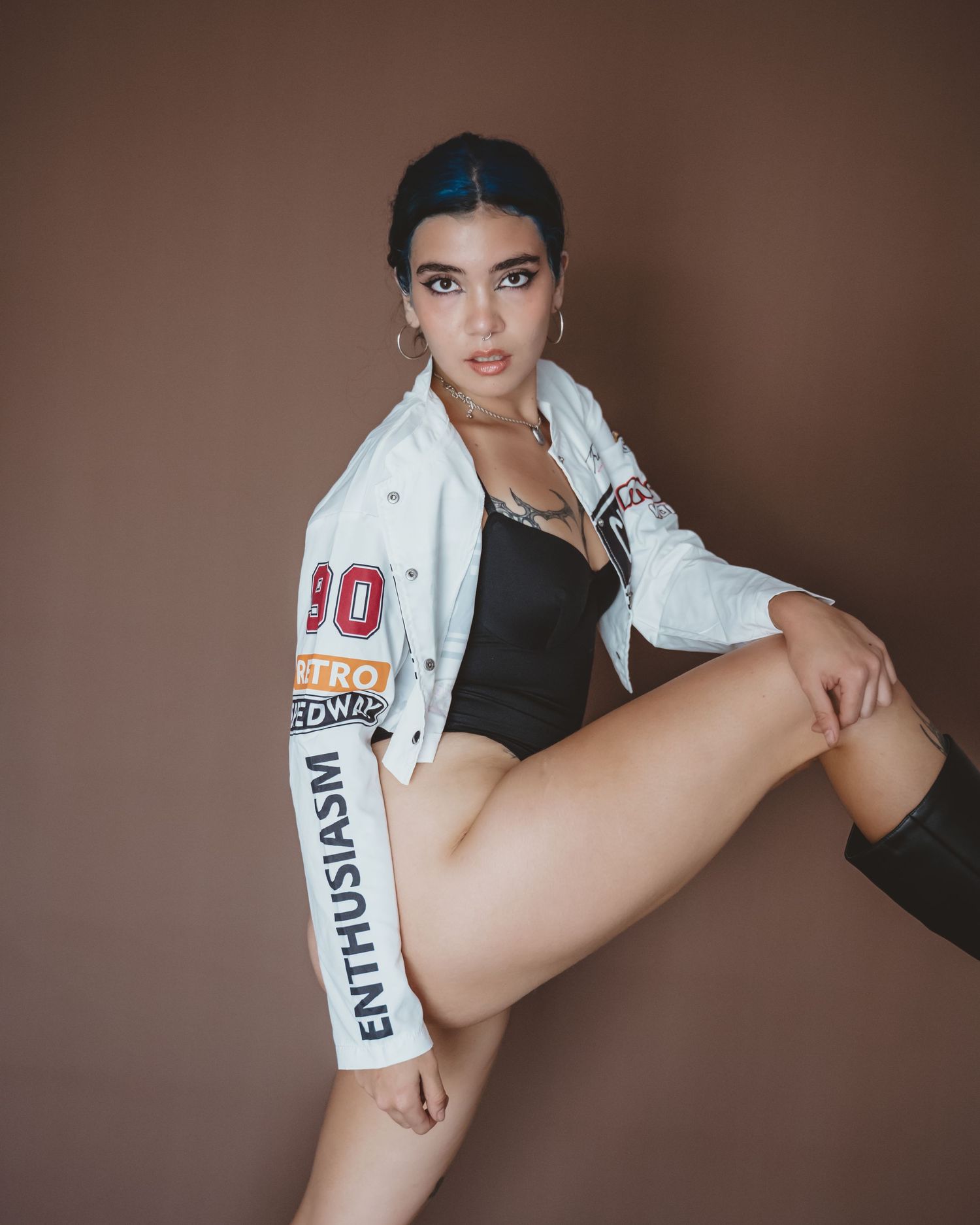 Woman with blue-streaked hair in a white graphic jacket and black bodysuit, raising one knee in a studio pose.