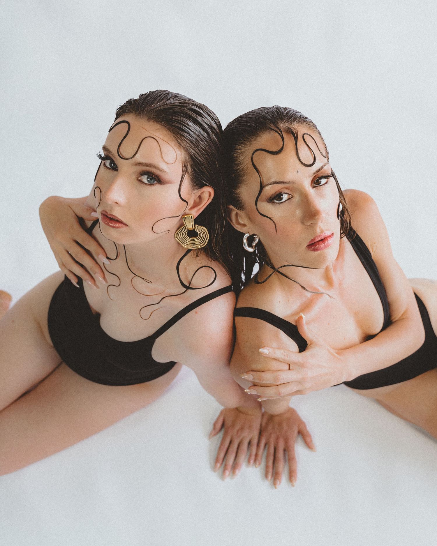 Two women in black swimsuits sit back to back on a white backdrop, with wavy painted lines on their faces and gold earrings.