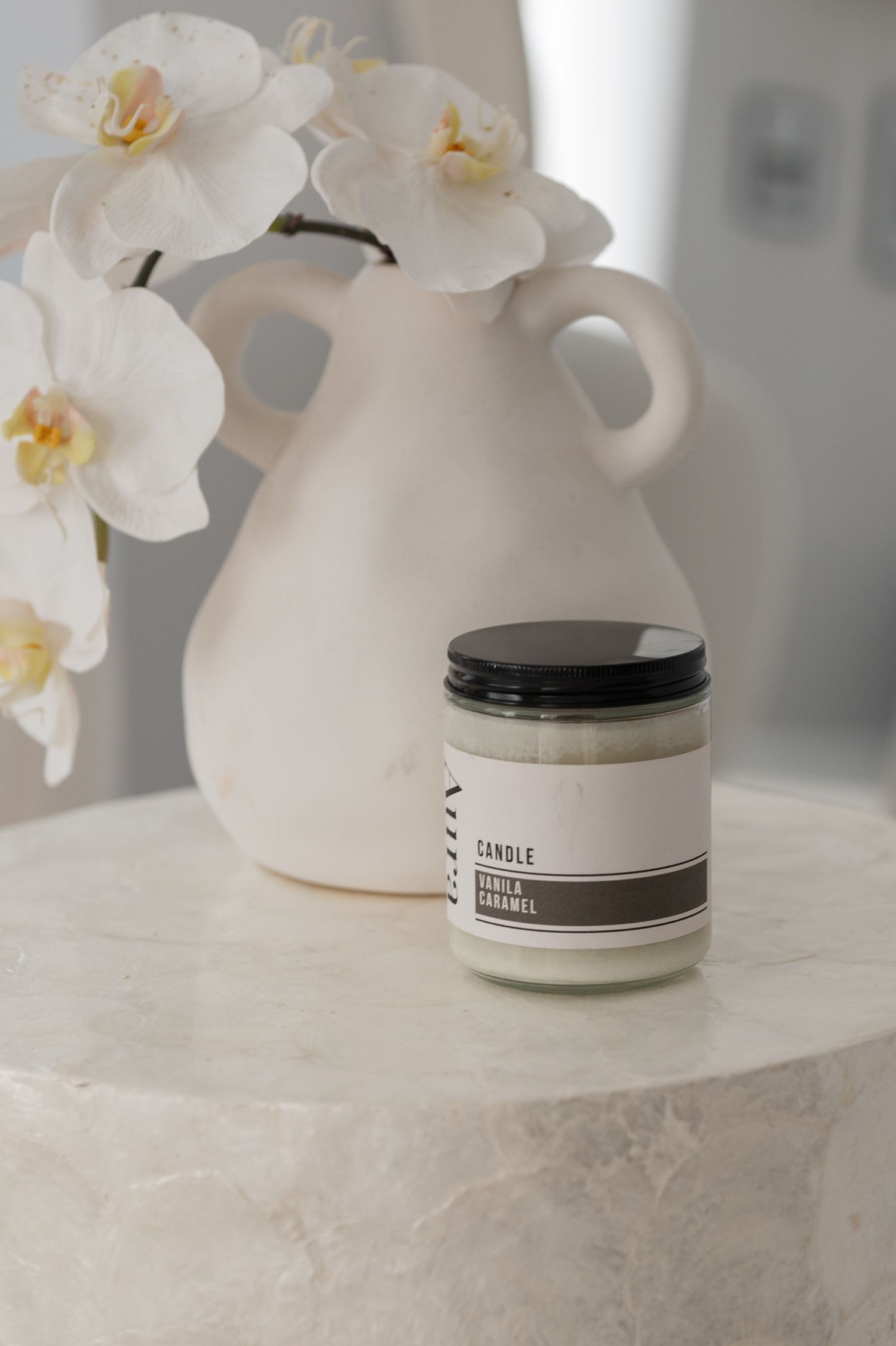 Vanilla caramel candle in a glass jar beside white orchids in a cream handled vase on a marble side table.