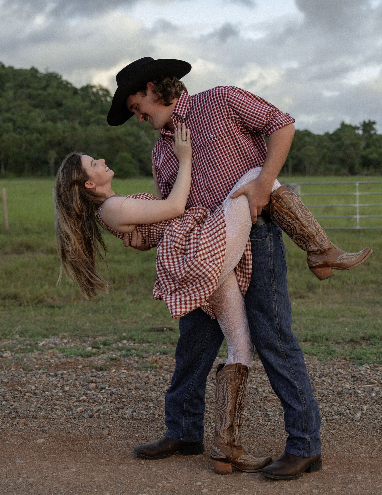 Cowboy in a black hat and jeans dips a woman in a gingham dress backward in a romantic country pose on a dirt road.