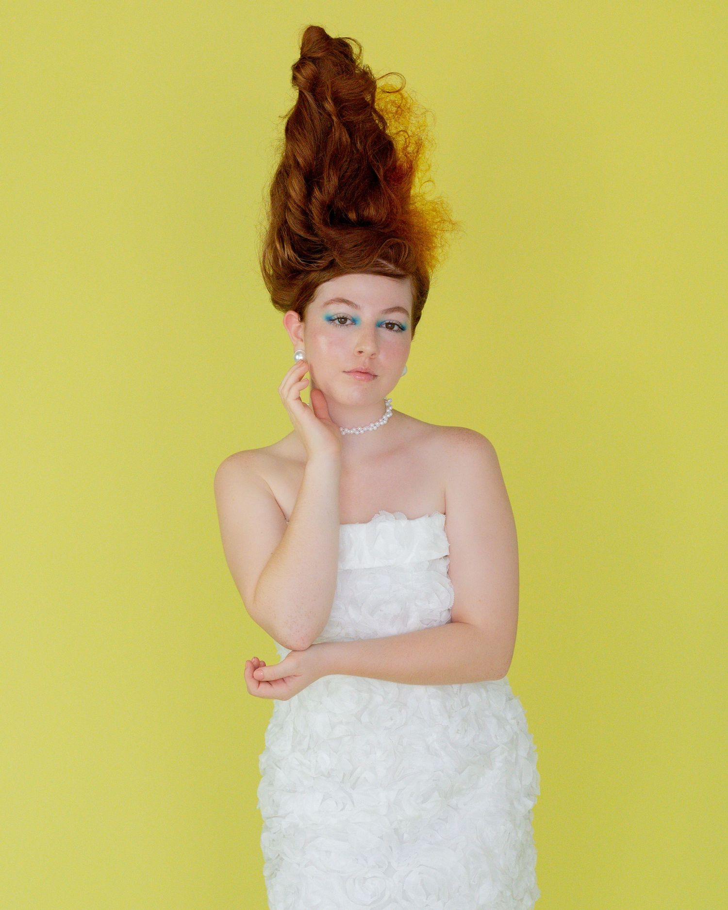 Model with a tall, sculpted auburn updo and blue eyeshadow wearing a white rosette strapless dress and pearl necklace on a yellow backdrop.
