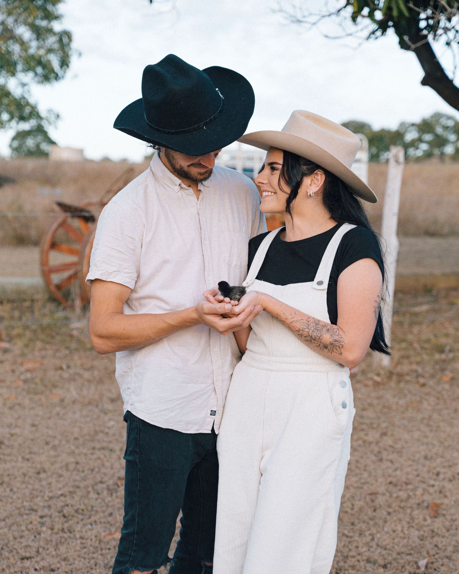 Smiling couple in cowboy hats stand close together holding a small object in a rustic country setting with an old wagon wheel.