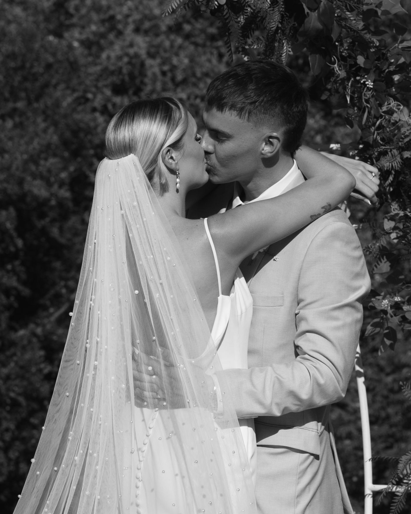 Black and white image of a bride with a pearl-studded veil kissing her groom beside a leafy wedding arch.