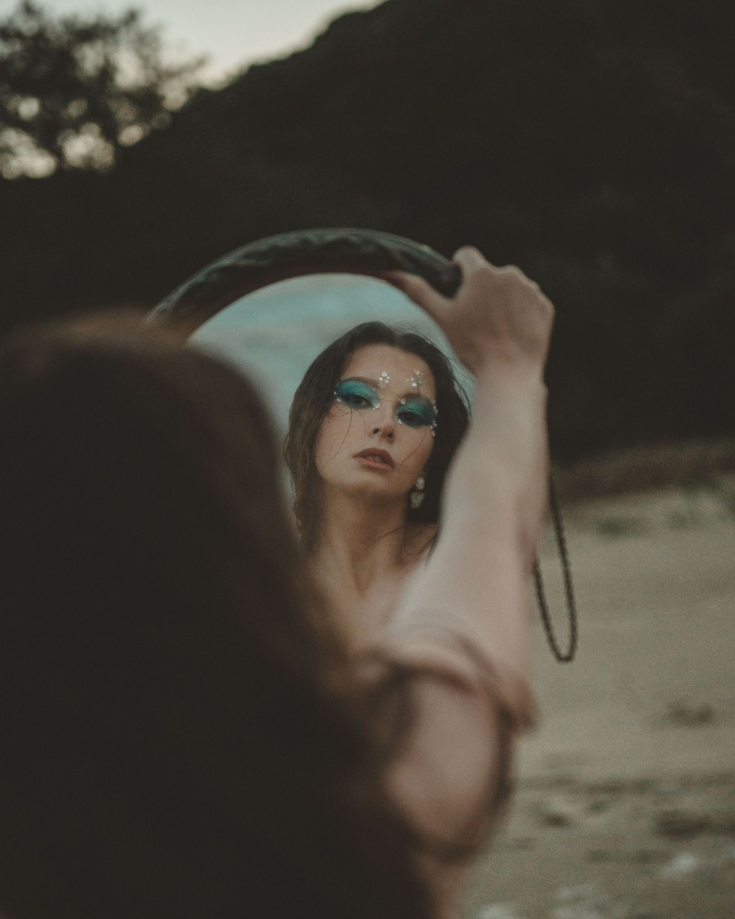 Woman with teal eye makeup and face gems gazes into a round mirror held up on a moody, overcast beach.
