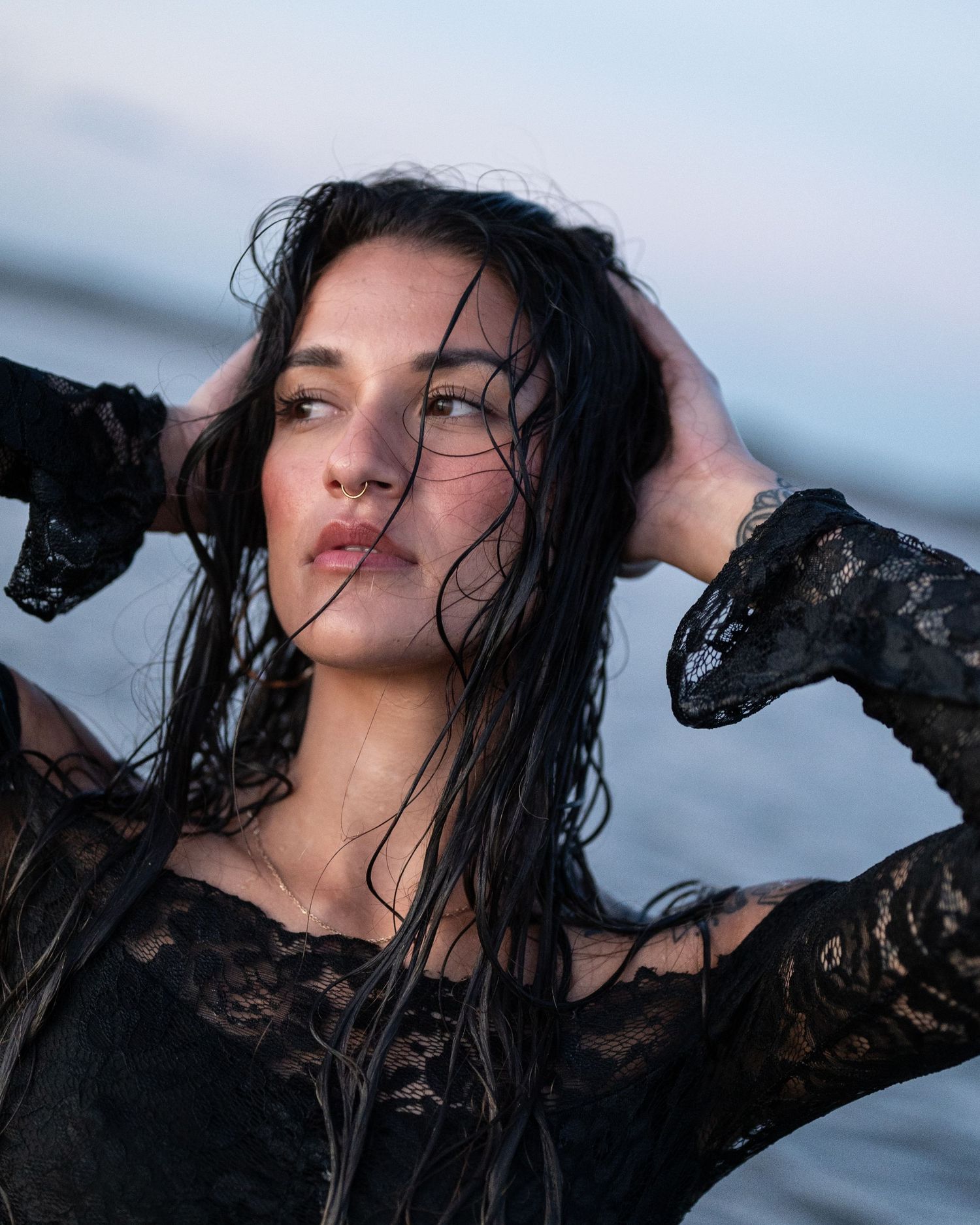 Close-up of a woman in black lace with wet, wind-swept hair, hands in her hair, by the sea at dusk.