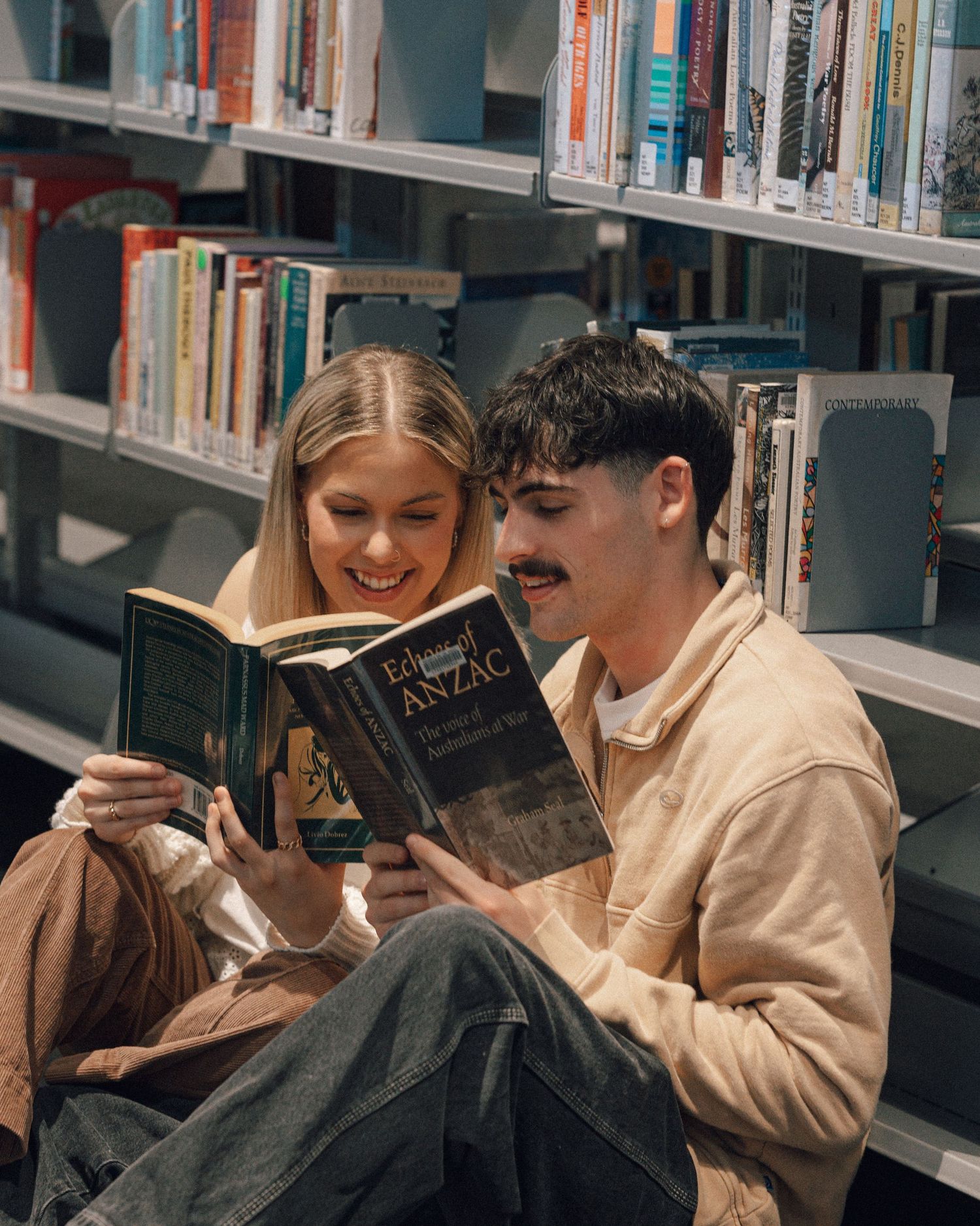 Smiling couple sit on the floor of a library reading a book together beside shelves filled with books.
