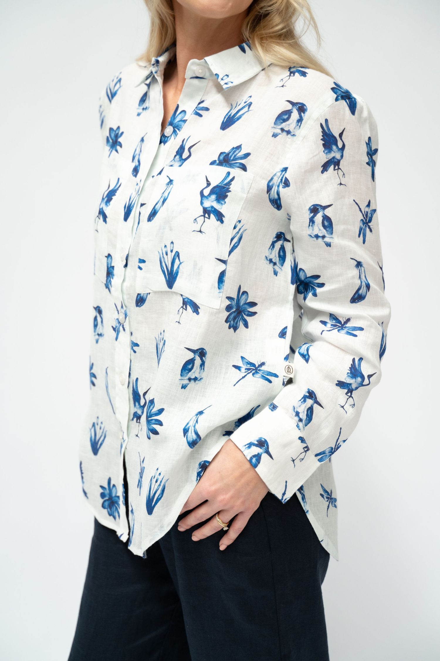 A woman wears a white linen button-up shirt with a blue bird and floral print, paired with navy pants, against a plain white background.
