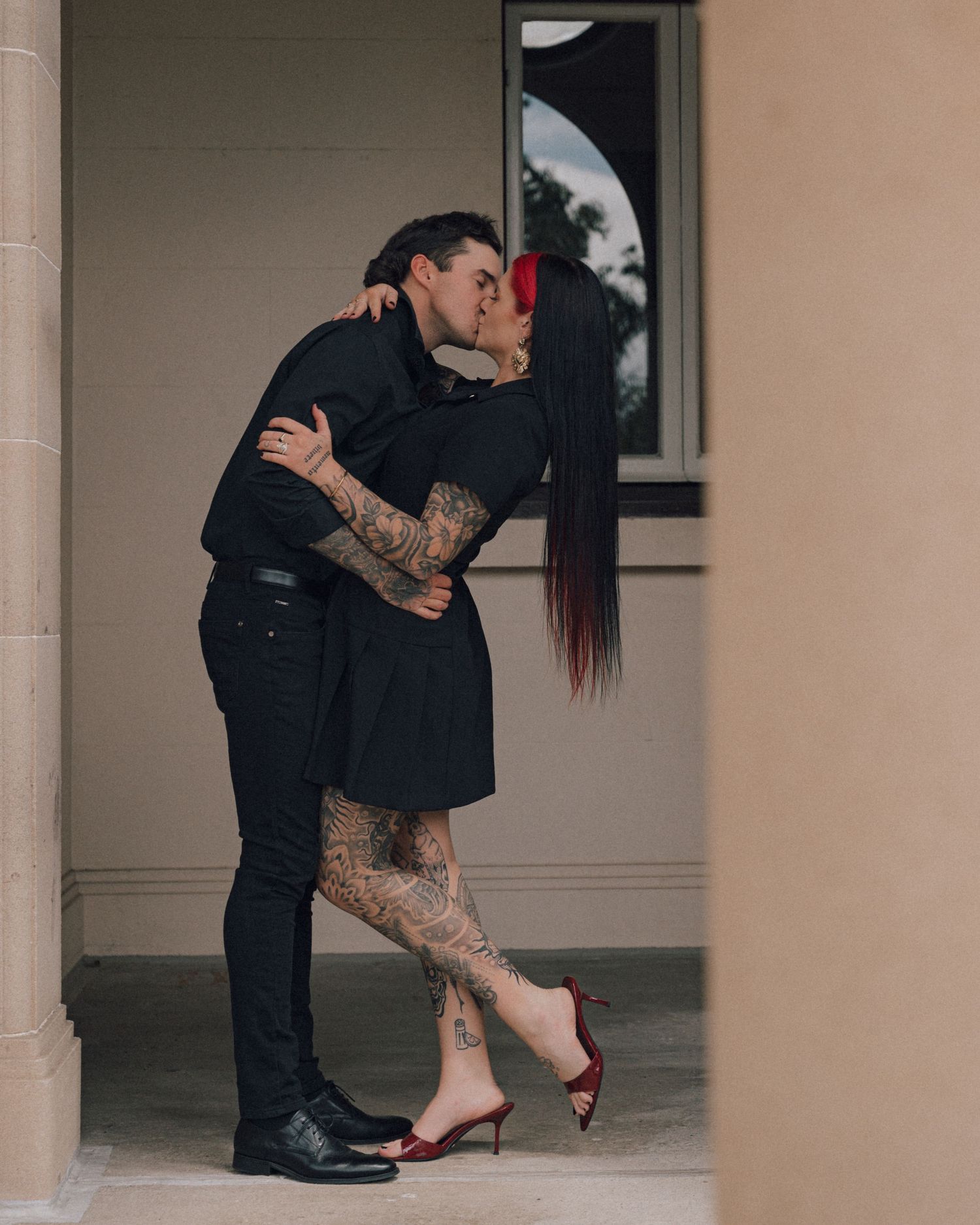 A tattooed couple in black outfits shares a kiss beside a stone column, the woman wearing red heels and a black dress.