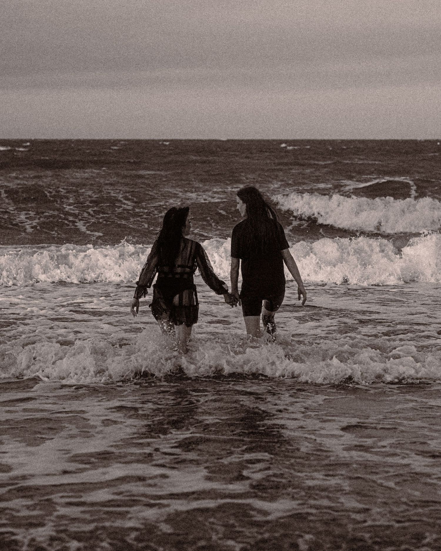 Black and white photo of two people holding hands as they wade into rolling ocean waves under a cloudy sky.