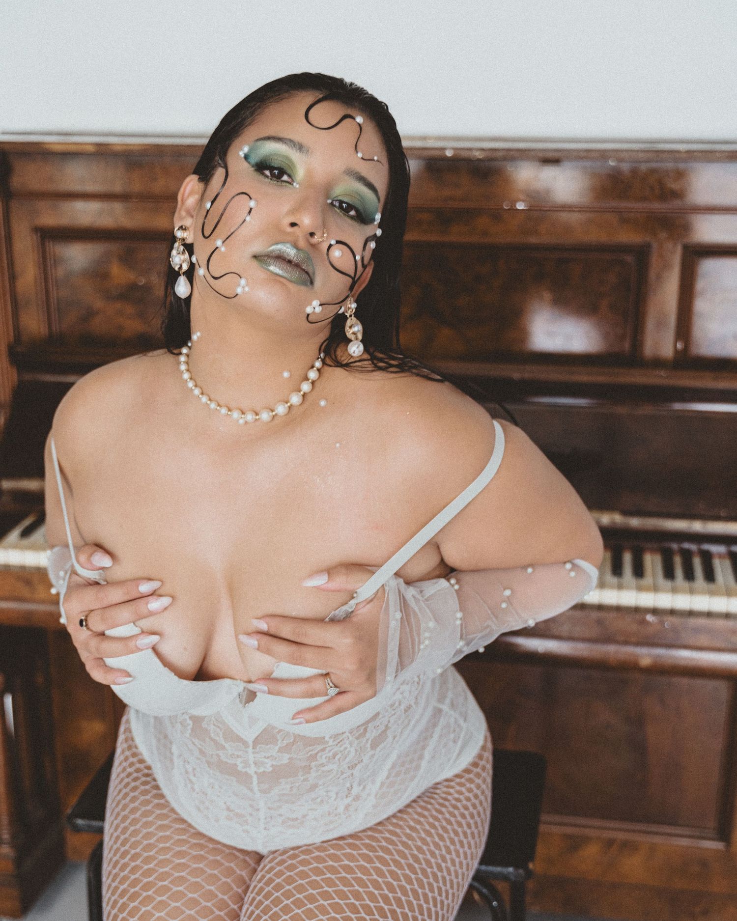 Woman in white lace bodysuit and fishnets sits at a piano, wearing pearl jewelry, green eye makeup and wire face art.