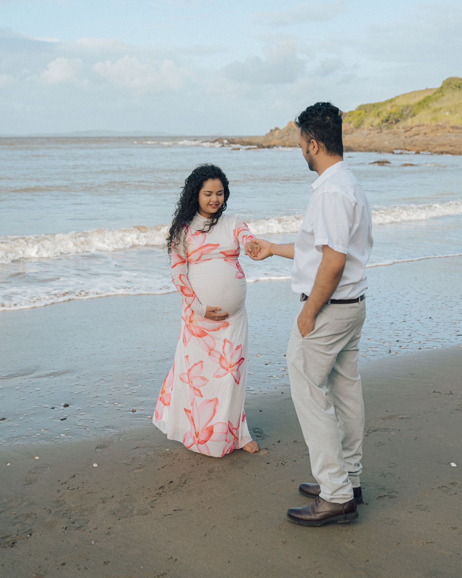 Expecting couple stands facing each other on the beach, the pregnant woman in a floral dress and the man in a white shirt and khakis.