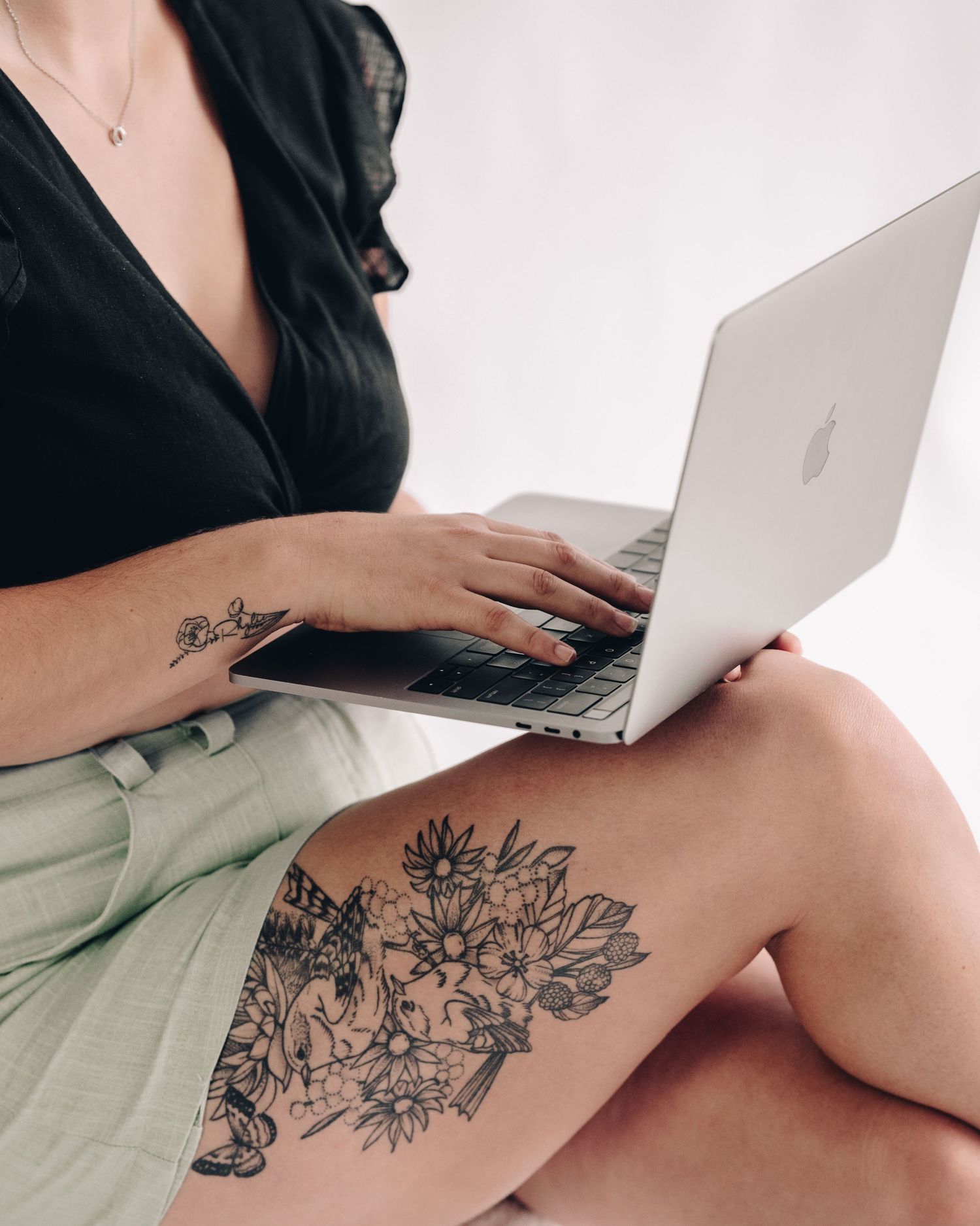 Woman with floral leg and arm tattoos in a black top and green shorts types on a laptop resting on her knees.