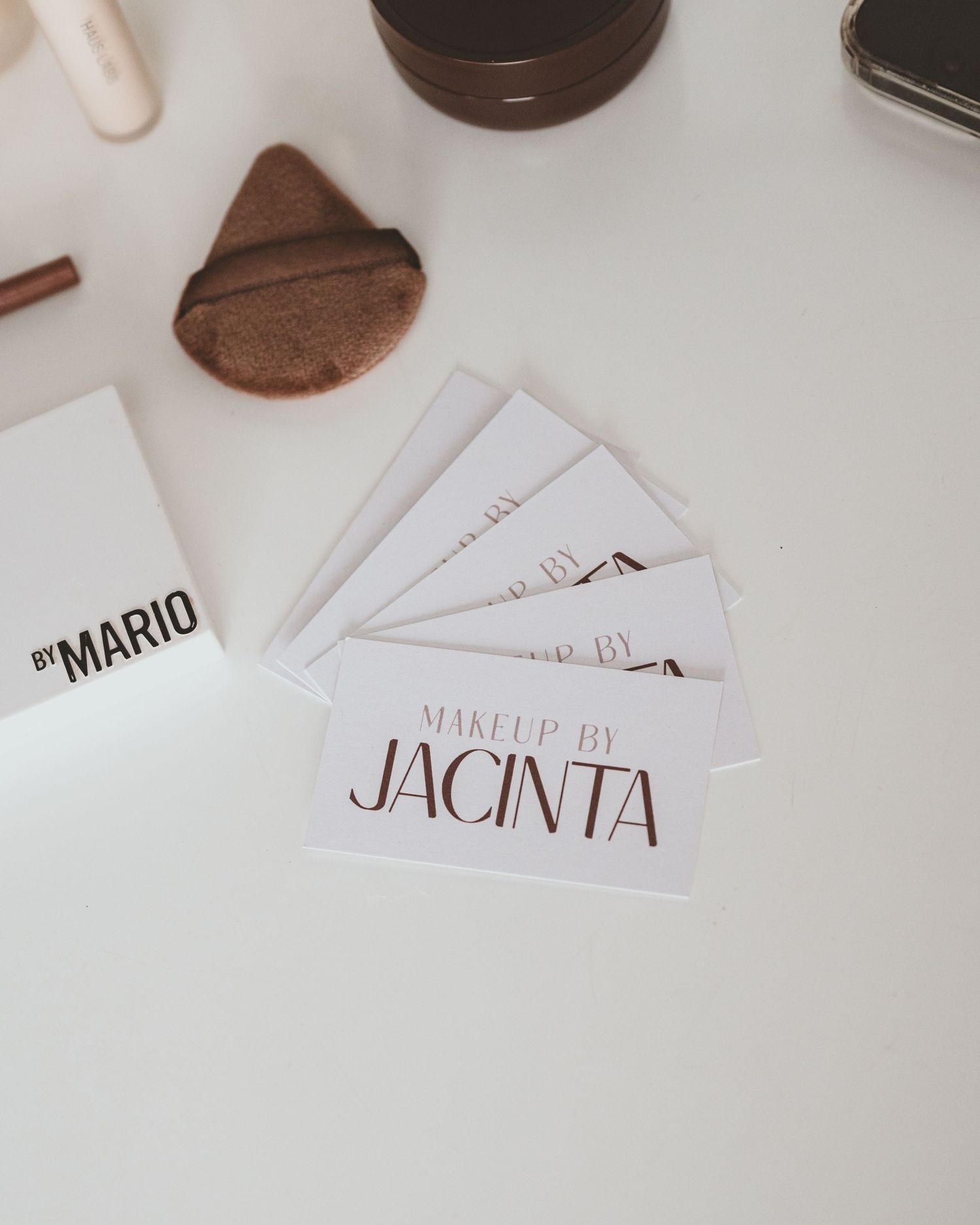 Makeup by Jacinta business cards fanned out on a white table beside a makeup sponge and a By Mario palette.